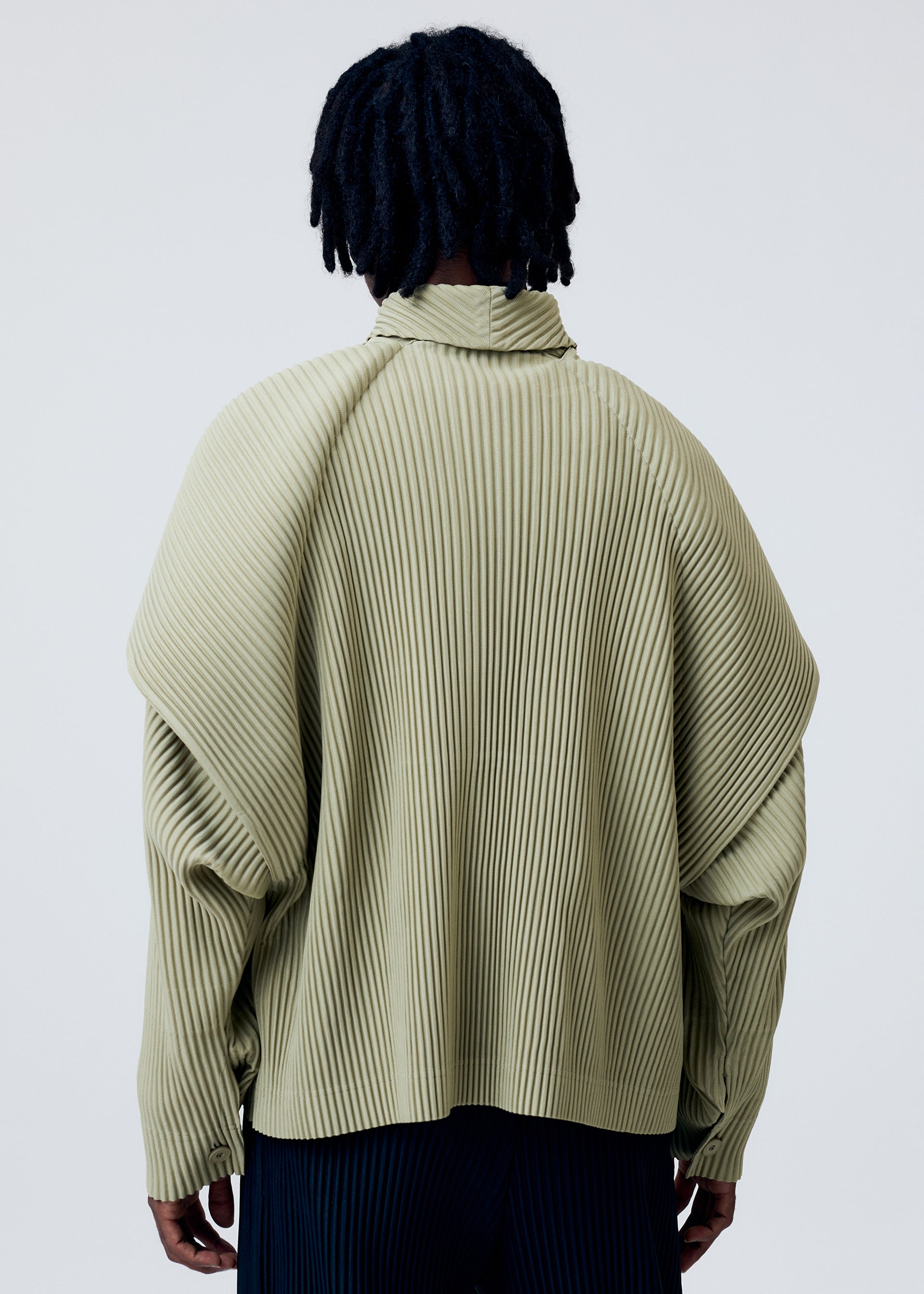 Zoomed view A person with short, textured black hair is seen from the back wearing the HOMME PLISSÉ ISSEY MIYAKE COVER AND FOLD top in light olive green, featuring wide shoulders, a high collar, and architectural 3D pleating against a plain light gray background.
