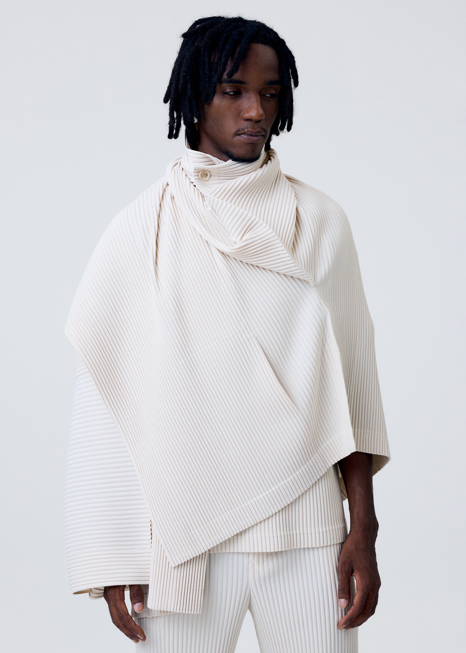 Zoomed view A person with medium-length dreadlocks wears the cream WEAR OR WRAP pleated asymmetrical poncho by HOMME PLISSÉ ISSEY MIYAKE over matching pants, standing against a plain light background.