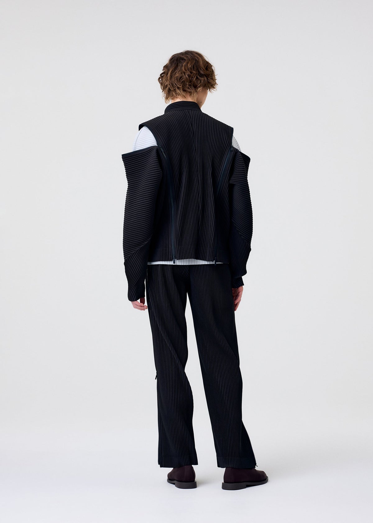 A person with wavy brown hair faces away, wearing the HOMME PLISSÉ ISSEY MIYAKE MULTI-FASTENED dark pinstriped suit with a uniquely structured jacket, suggesting versatile styling. The background is plain and light-colored. view 9 of 11