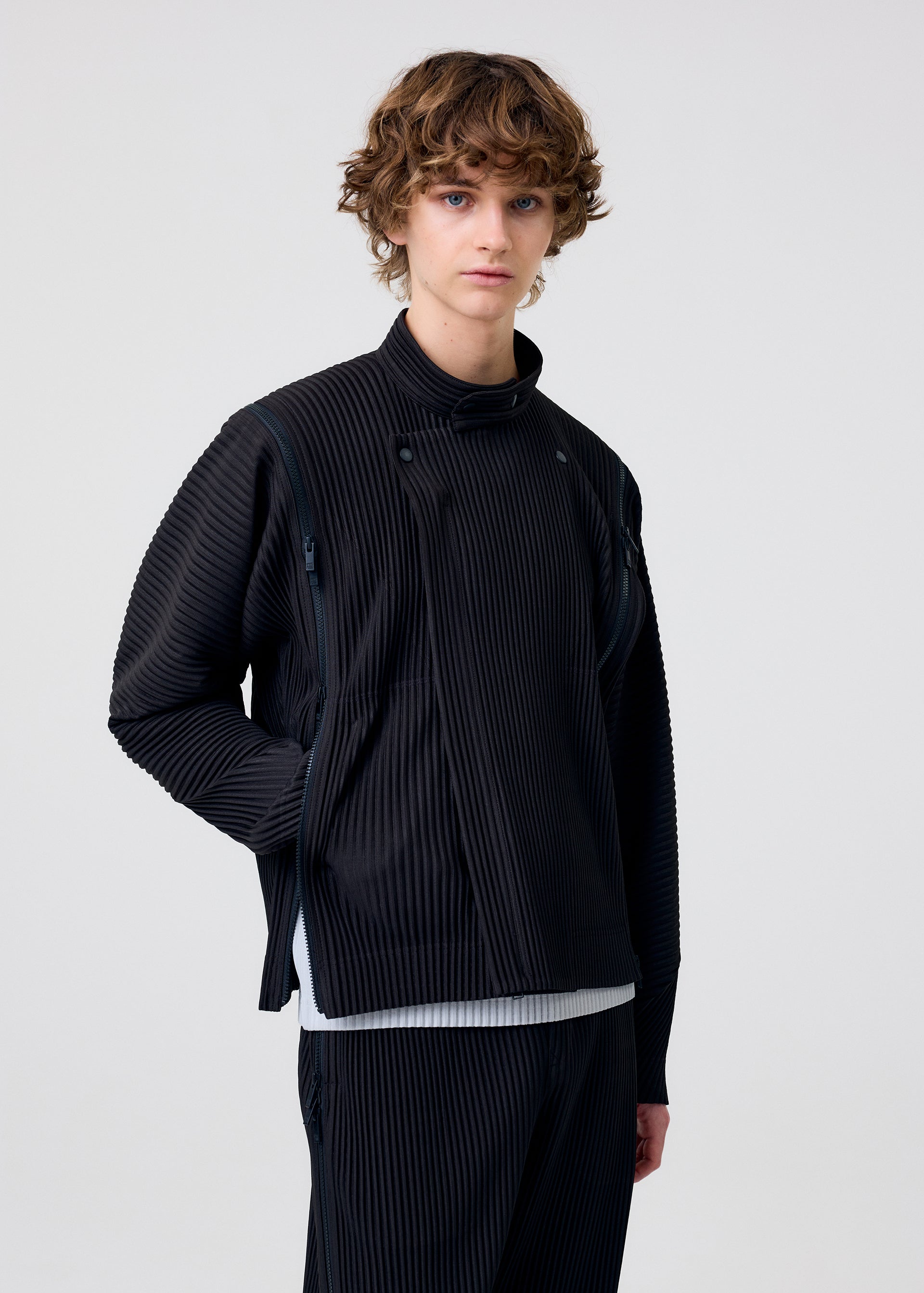 Zoomed view A person with curly brown hair stands against a plain background, wearing the HOMME PLISSÉ ISSEY MIYAKE MULTI-FASTENED black jacket over a white shirt and matching black pants, looking directly at the camera with a neutral expression.