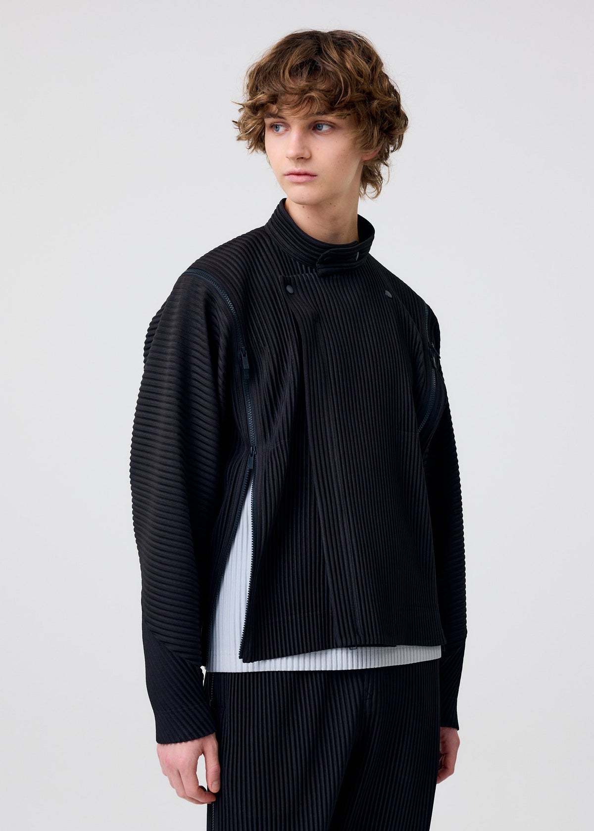 A person with wavy brown hair models the MULTI-FASTENED pleated black long-sleeve top and matching pants by HOMME PLISSÉ ISSEY MIYAKE, styled over a white shirt against a plain light background, gazing off to the side. view 11 of 11