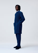 A man stands in profile against a plain light background, wearing HOMME PLISSÉ ISSEY MIYAKE's TAILORED PLEATS 1—a matching blue pinstriped collarless long jacket and trousers—effortlessly bridging formal and casual style.