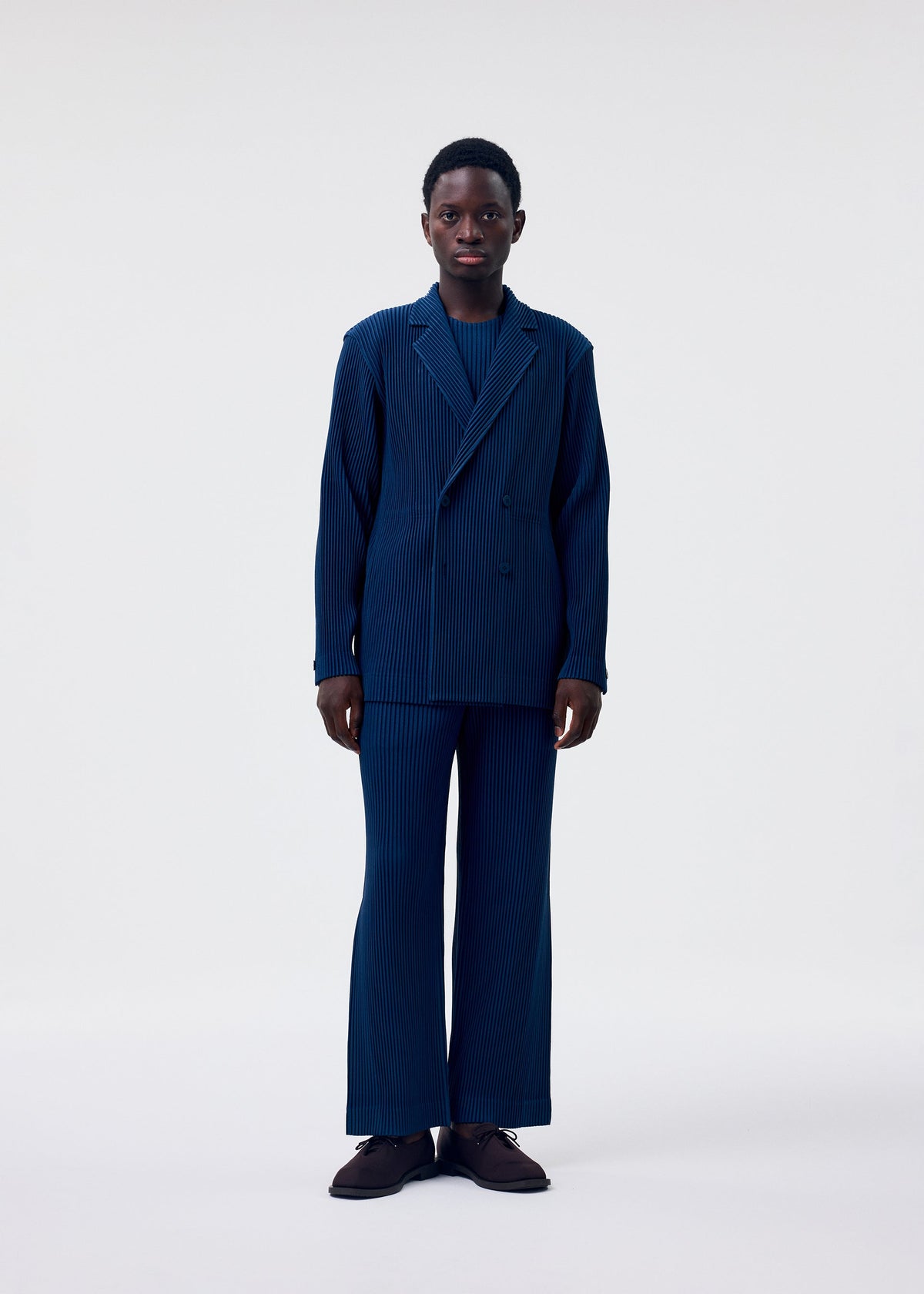 A person stands facing forward against a plain white background, wearing the TAILORED PLEATS 1 blue pinstripe suit by HOMME PLISSÉ ISSEY MIYAKE, featuring wide-leg pants, a double-breasted jacket, and dark shoes. view 6 of 7