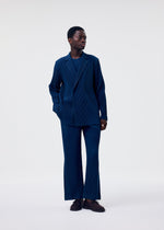 A person stands against a plain background wearing the HOMME PLISSÉ ISSEY MIYAKE TAILORED PLEATS 1 suit—dark blue pinstripes with matching trousers, shirt, and dark shoes. One hand is in their pocket as they look to the side.