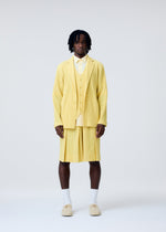 A man stands against a plain background wearing HOMME PLISSÉ ISSEY MIYAKE’s TAILORED PLEATS 2—a light yellow quick-drying polyester jacket and pleated shorts—paired with a yellow shirt, white socks, and beige shoes. His short dreadlocks frame a neutral expression.