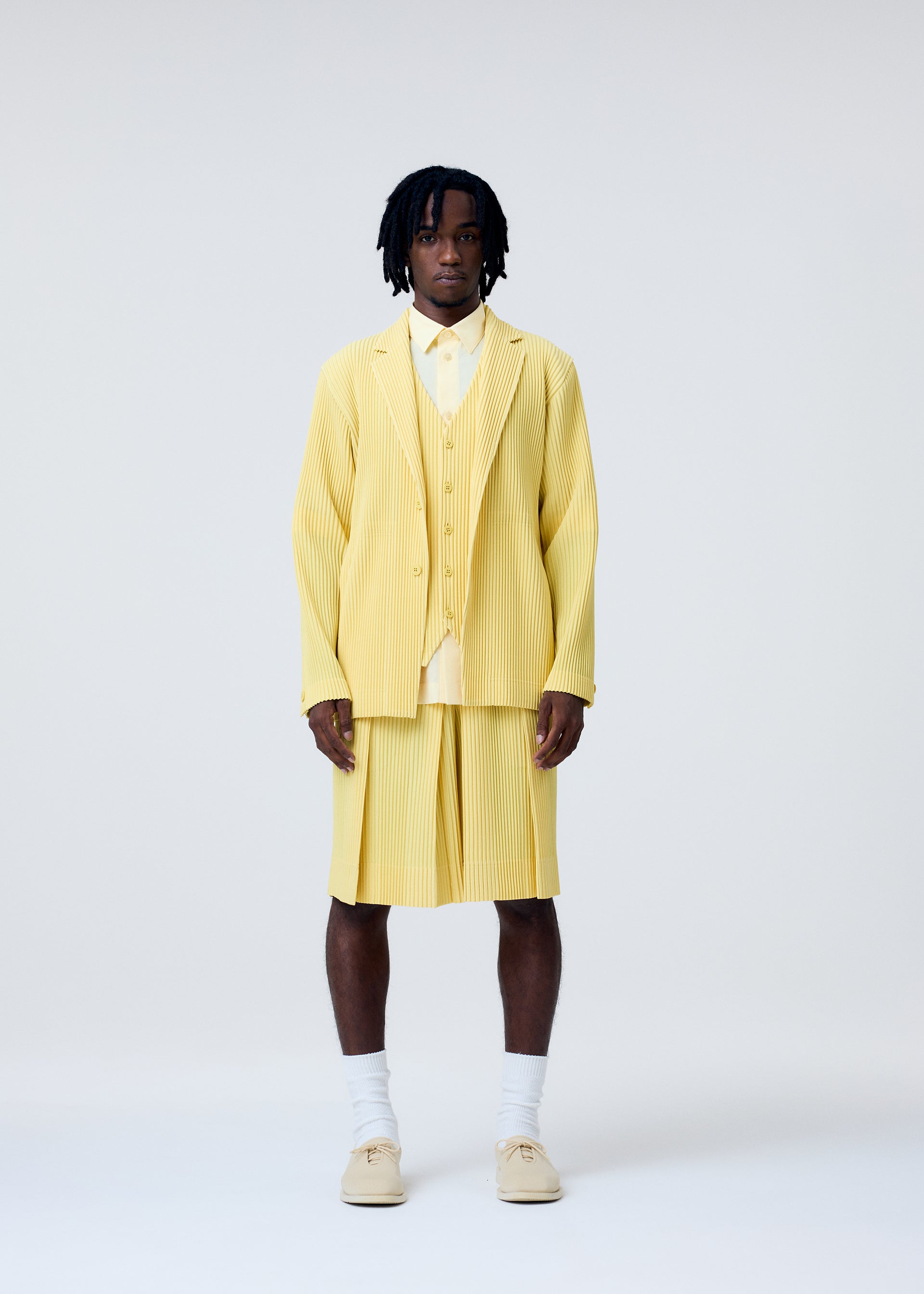 Zoomed view A man stands against a plain background wearing HOMME PLISSÉ ISSEY MIYAKE’s TAILORED PLEATS 2—a light yellow quick-drying polyester jacket and pleated shorts—paired with a yellow shirt, white socks, and beige shoes. His short dreadlocks frame a neutral expression.