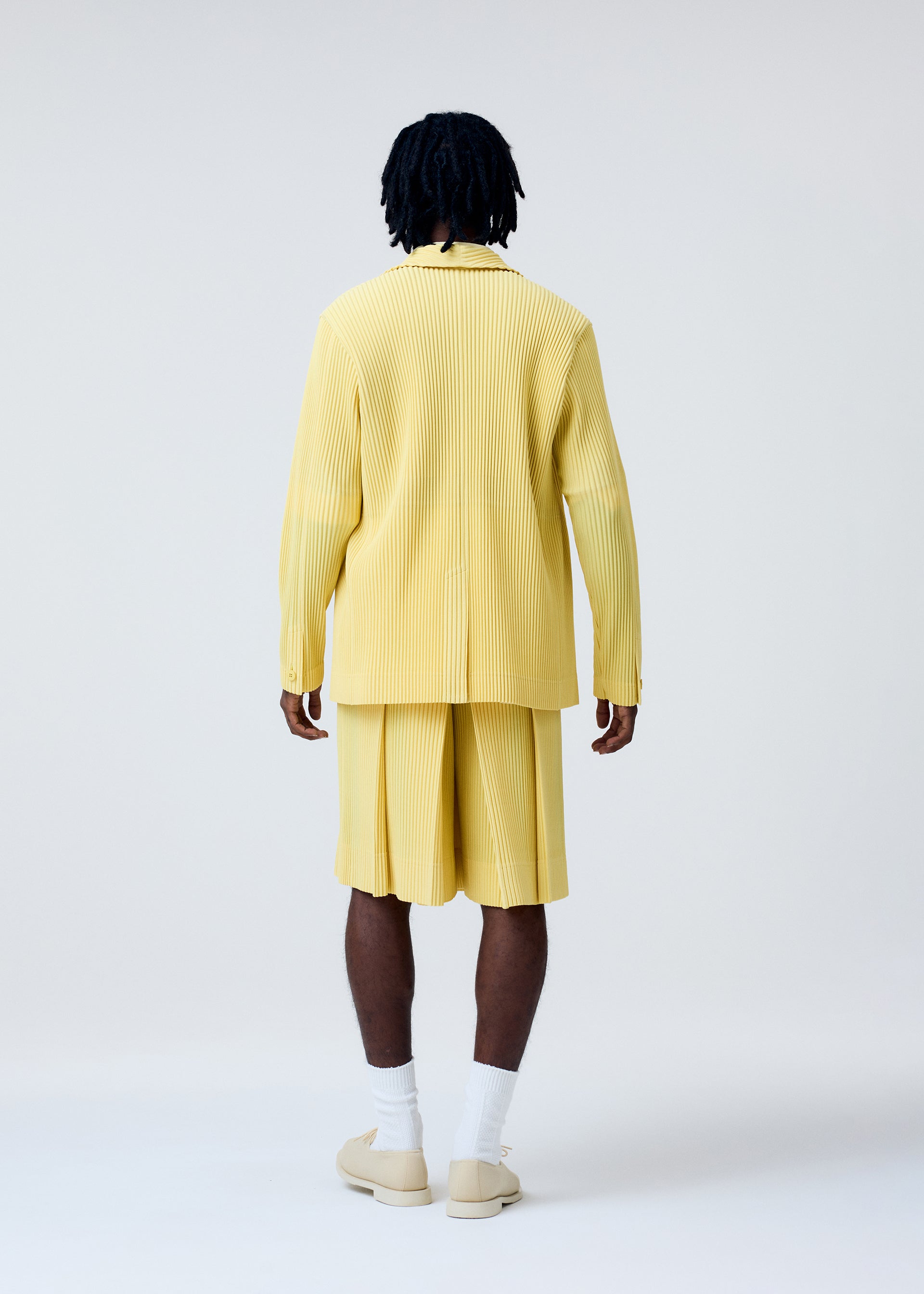 Zoomed view A person with short dreadlocks faces away, wearing HOMME PLISSÉ ISSEY MIYAKE's TAILORED PLEATS 2 yellow polyester jacket and knee-length shorts, with white socks and beige slip-ons, set against a plain light background.