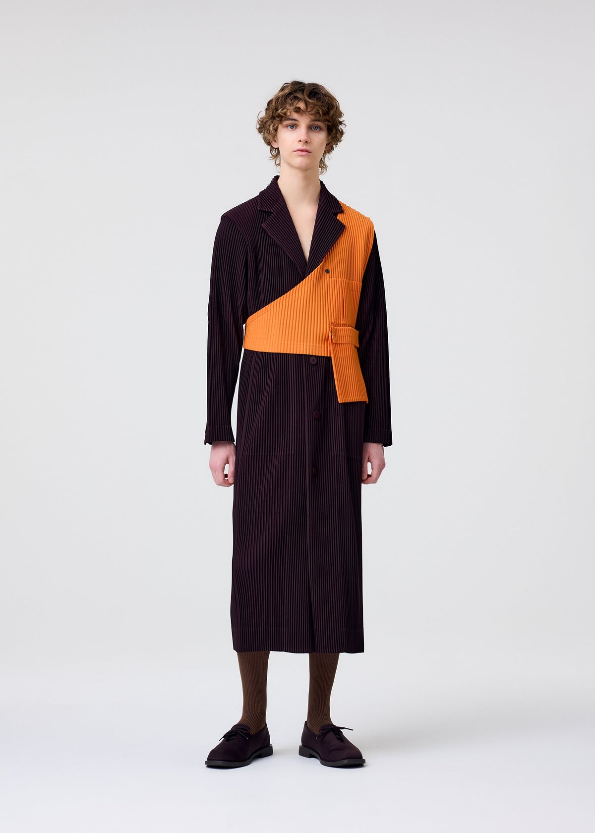 A person with curly hair faces forward against a plain background, wearing HOMME PLISSÉ ISSEY MIYAKE's TAILORED PLEATS 2: a long, dark pinstriped jacket with an asymmetrical orange panel, paired with brown socks and dark shoes. view 5 of 6