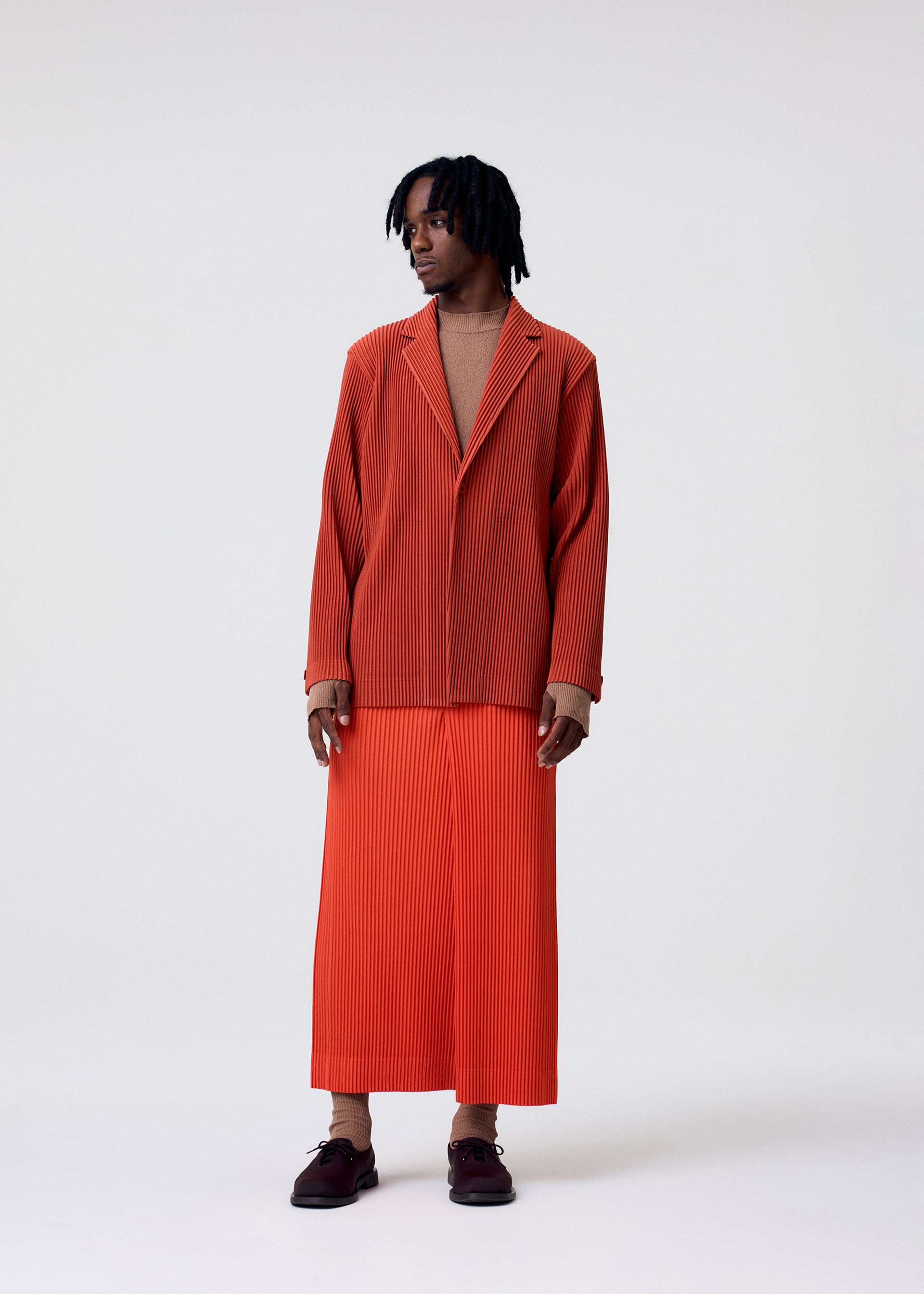 Zoomed view A person with short locs wears HOMME PLISSÉ ISSEY MIYAKE TAILORED PLEATS 3: a loose, pleated burnt orange blazer and matching long skirt over a beige top, with brown shoes and socks, posed against a plain light background.