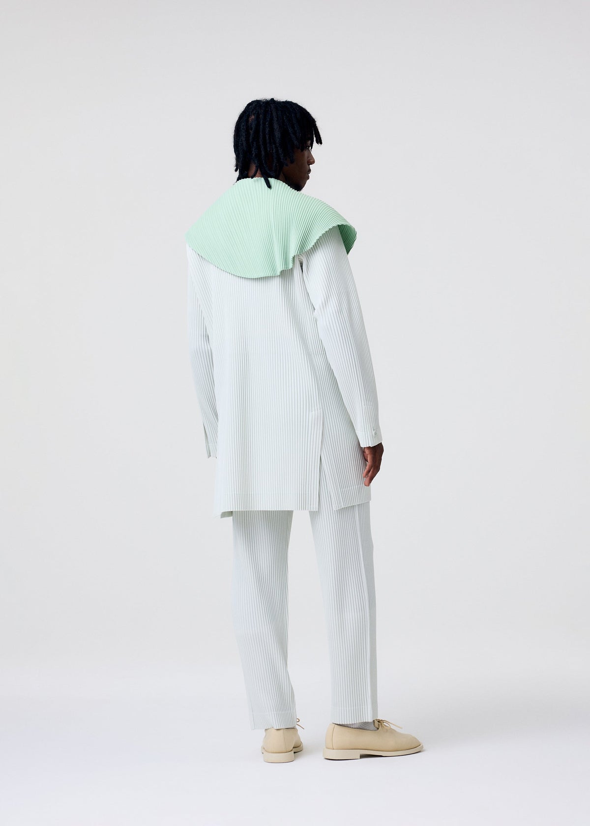 A person with short, dark, textured hair stands facing away in a light pinstriped TAILORED PLEATS 3 set by HOMME PLISSÉ ISSEY MIYAKE, mint green shawl over their shoulders, beige shoes, against a plain white background. view 6 of 6