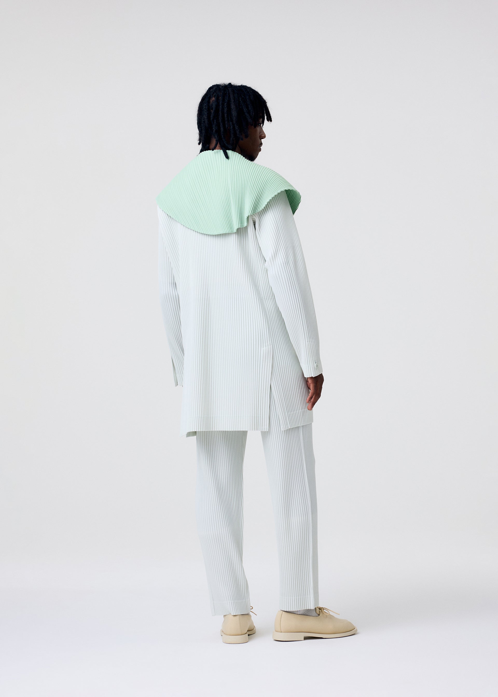 Zoomed view A person with short, dark, textured hair stands facing away in a light pinstriped TAILORED PLEATS 3 set by HOMME PLISSÉ ISSEY MIYAKE, mint green shawl over their shoulders, beige shoes, against a plain white background.