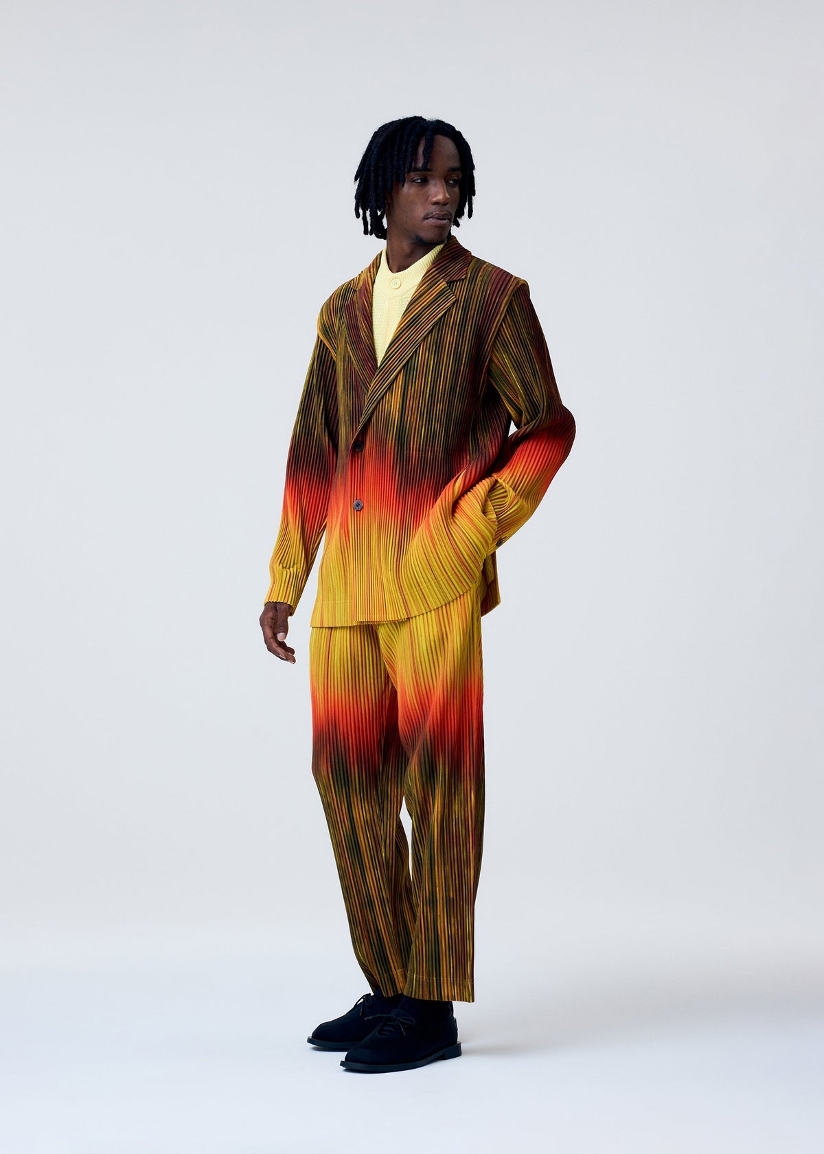 A person poses against a plain background wearing the HOMME PLISSÉ ISSEY MIYAKE PAINT BRUSH CLOSE-UP jacket in yellow, orange, and red over a yellow shirt with black shoes, glancing sideways with one hand in their pocket. view 6 of 8