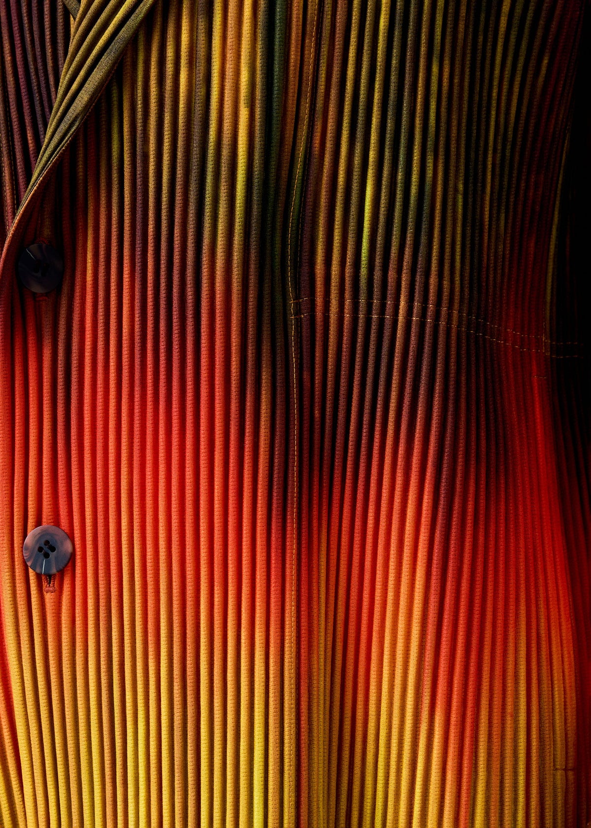 Close-up of the PAINT BRUSH CLOSE-UP jacket from HOMME PLISSÉ ISSEY MIYAKE, featuring pleated fabric with a gradient from dark green to yellow, orange, and bright red, and a double-breasted design with two dark buttons on the left. view 8 of 8