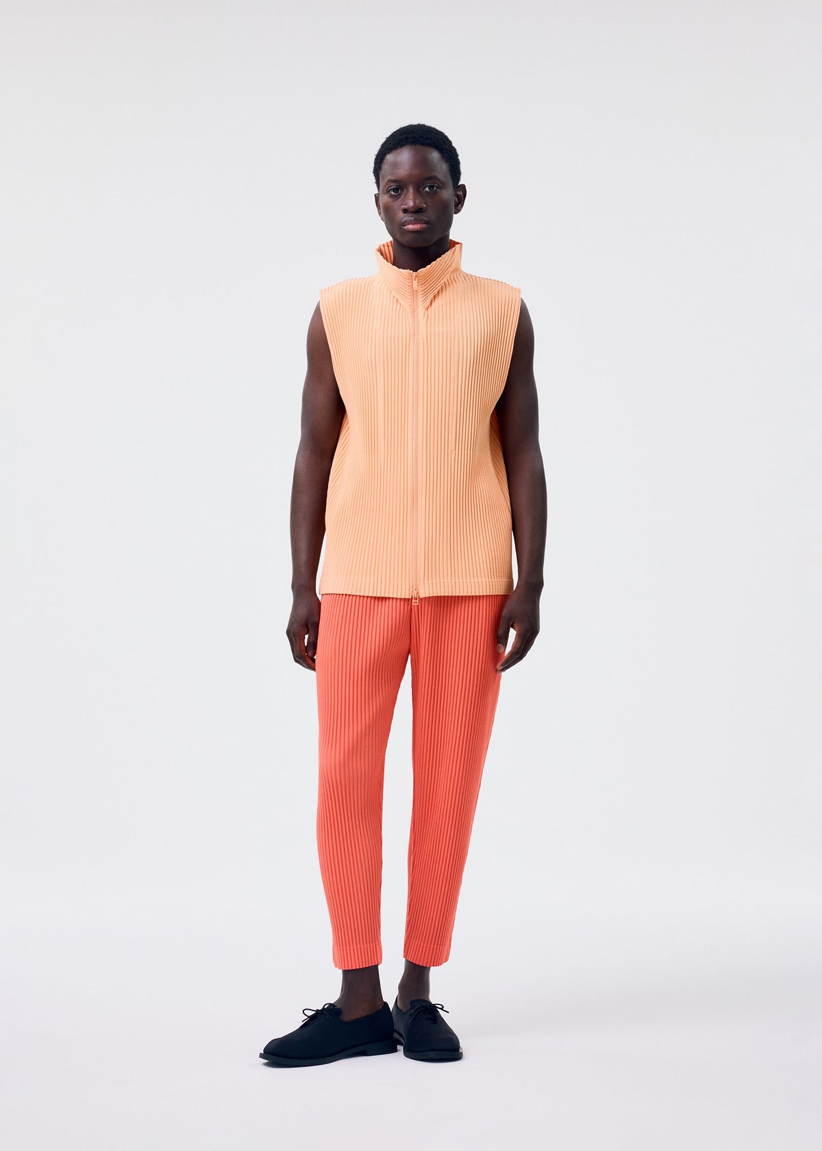 A man with short hair and black shoes stands against a plain background, wearing HOMME PLISSÉ ISSEY MIYAKE's MONTHLY COLORS : APRIL—a sleeveless light orange pleated top and orange pleated pants designed for comfort. view 7 of 7
