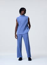 A person with short, textured hair stands against a light background, wearing HOMME PLISSÉ ISSEY MIYAKE's TAILORED PLEATS 1: a quick-drying sleeveless high-neck top and matching wide-leg blue pants, with black shoes and one hand in their pocket.