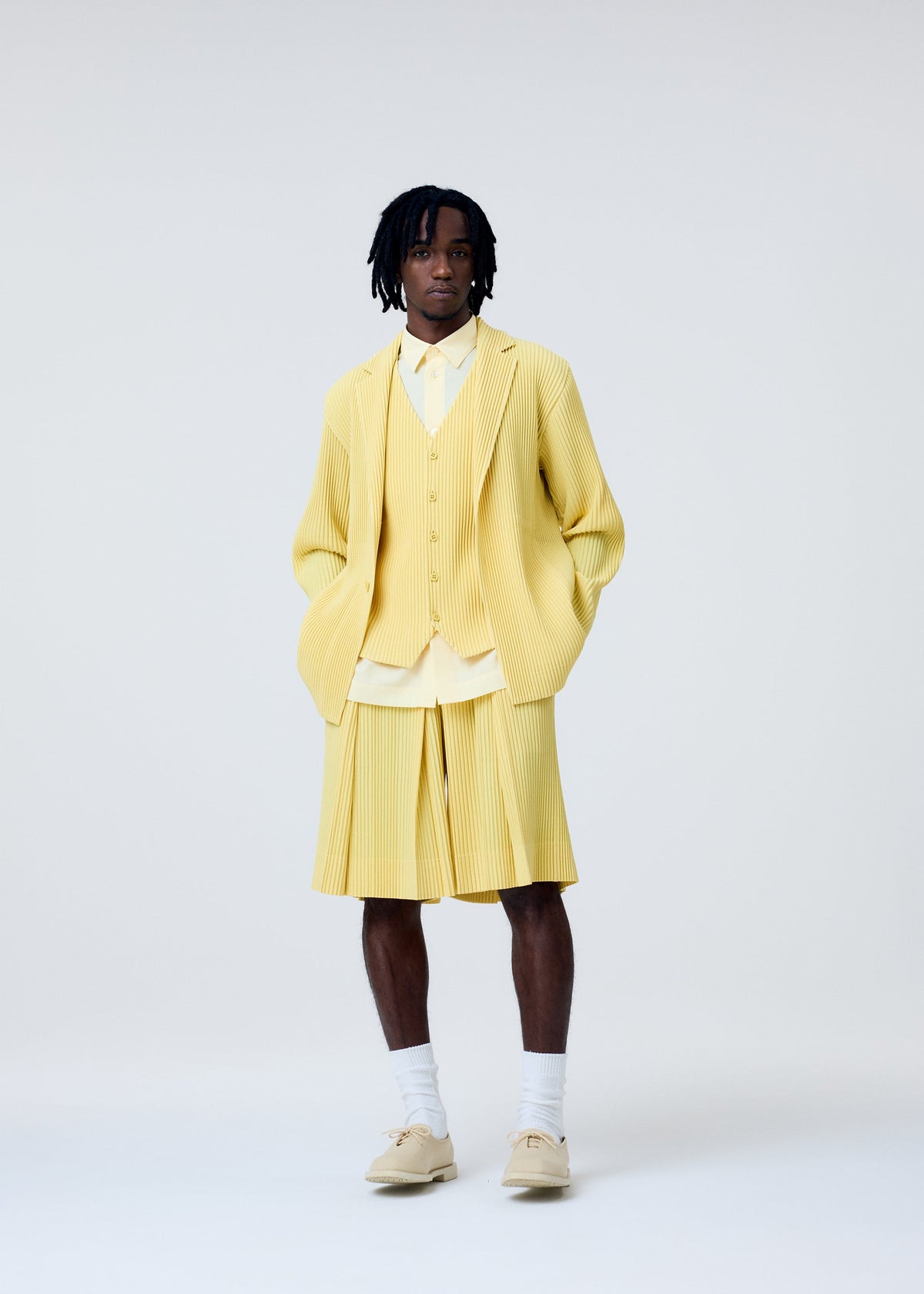 A person with short dreadlocks and a neutral expression wears the HOMME PLISSÉ ISSEY MIYAKE TAILORED PLEATS 2 suit—light yellow pleated jacket, matching shorts, quick-drying vest—styled with a light shirt, white socks, and beige shoes against a plain background. view 6 of 7