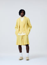 A person with short dreadlocks and a neutral expression wears the HOMME PLISSÉ ISSEY MIYAKE TAILORED PLEATS 2 suit—light yellow pleated jacket, matching shorts, quick-drying vest—styled with a light shirt, white socks, and beige shoes against a plain background.