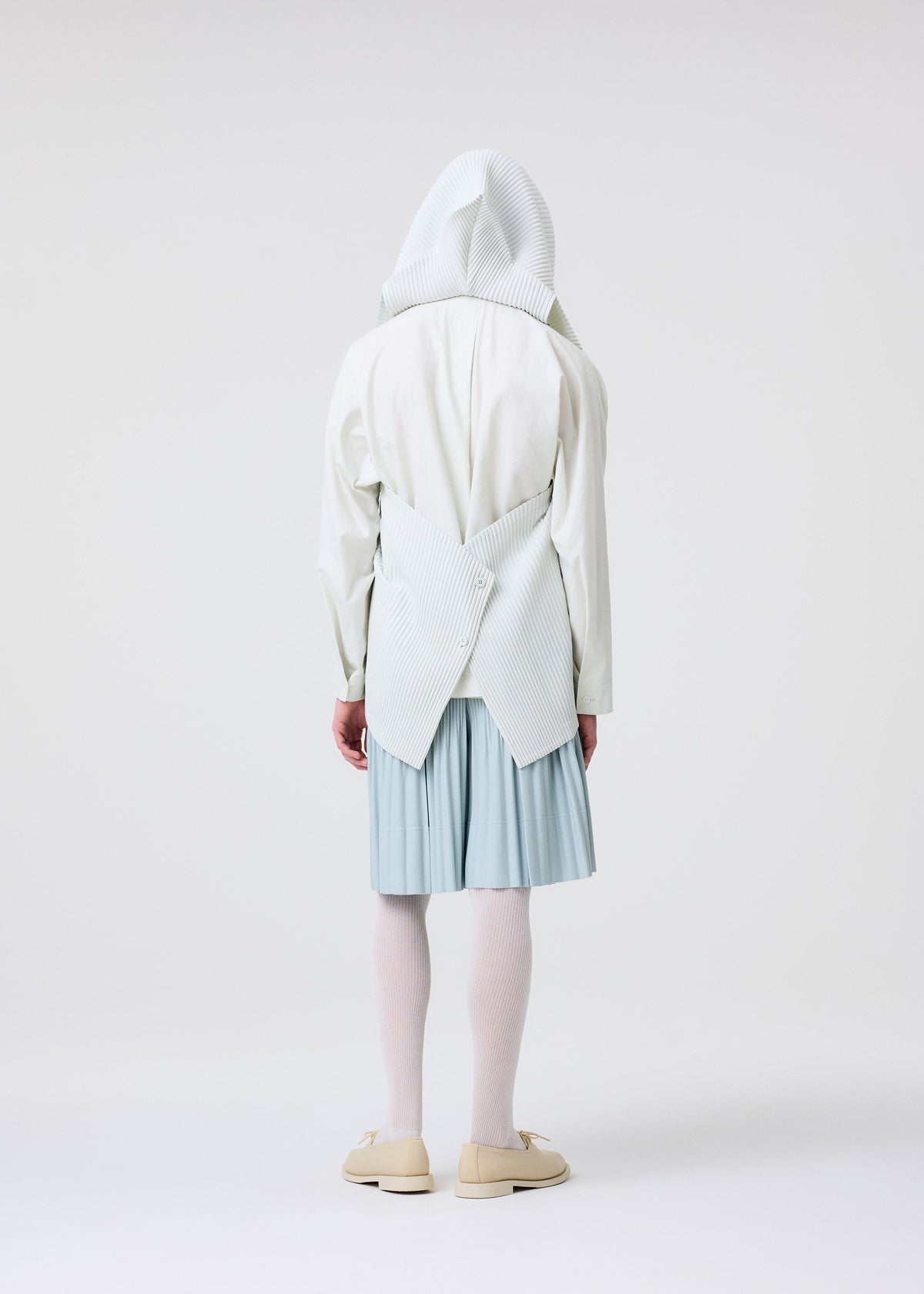 A person faces away, wearing the HOMME PLISSÉ ISSEY MIYAKE TAILORED PLEATS 3 white hooded top with a sculptural pleated back, paired with a light blue pleated skirt, white tights, and beige shoes against a plain white background. view 7 of 7
