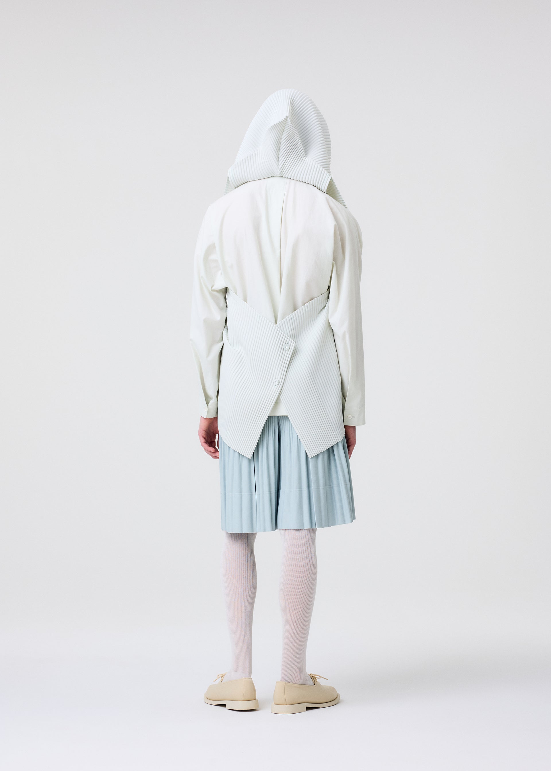 Zoomed view A person faces away, wearing the HOMME PLISSÉ ISSEY MIYAKE TAILORED PLEATS 3 white hooded top with a sculptural pleated back, paired with a light blue pleated skirt, white tights, and beige shoes against a plain white background.