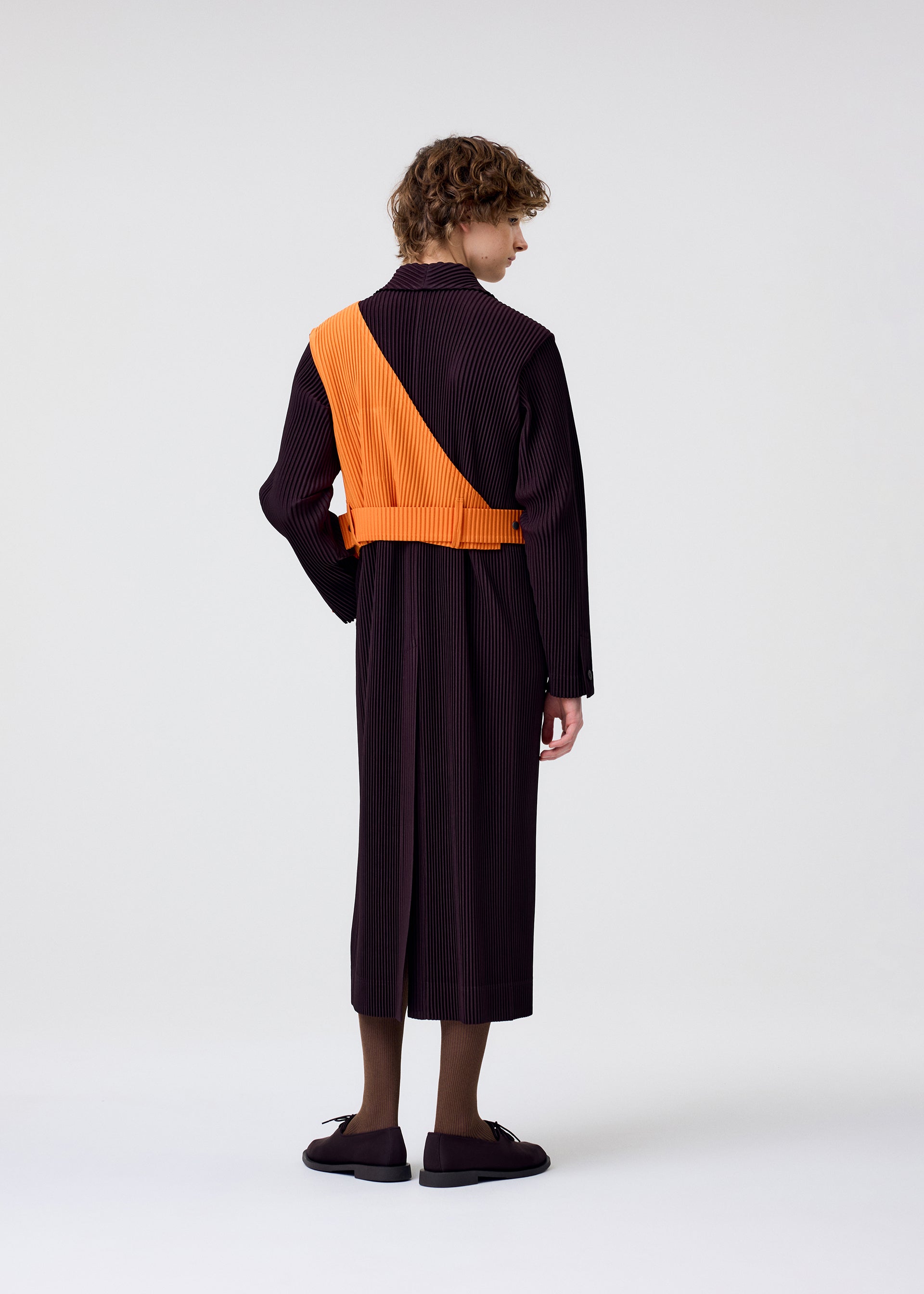 Zoomed view A person with curly hair faces away, wearing HOMME PLISSÉ ISSEY MIYAKE PAINTER’S GEAR: a long dark pinstriped coat with a bold orange one-shoulder vest draped diagonally, paired with dark shoes and semi-sheer brown socks, against a plain background.