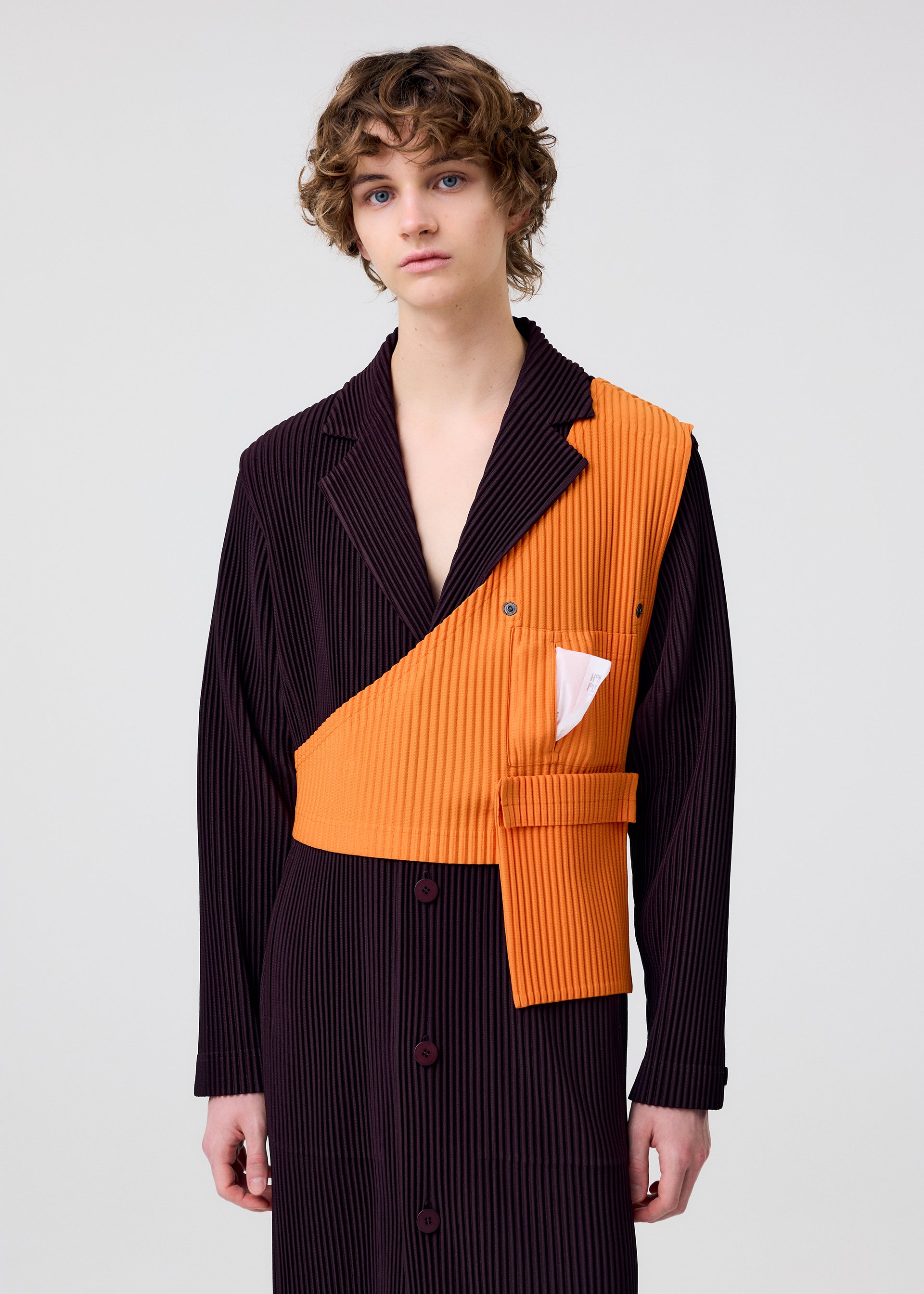 Zoomed view A person with short, curly hair wears HOMME PLISSÉ ISSEY MIYAKE PAINTER’S GEAR: a long pinstripe coat in dark and bright orange with an asymmetrical lapel, a white pocket handkerchief, and a versatile utility vest. The background is light gray.