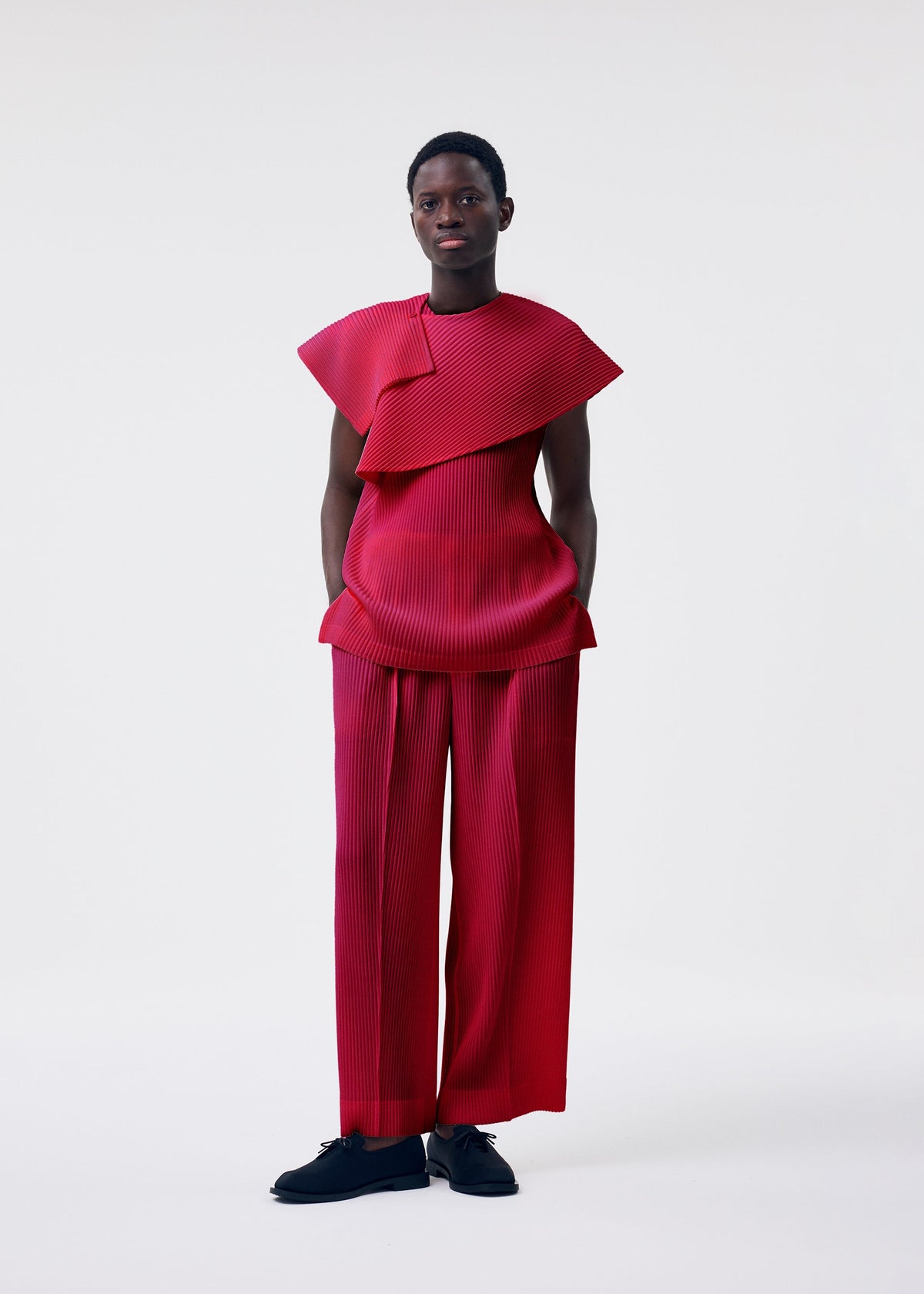 A person stands against a plain background wearing the HOMME PLISSÉ ISSEY MIYAKE MONTHLY COLORS : JANUARY set—a vibrant red, pleated, asymmetrical top with matching lightweight pants—paired with black shoes. view 6 of 7