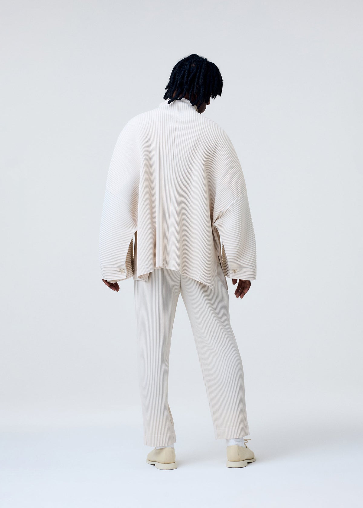 A person with dark, shoulder-length hair faces away, wearing HOMME PLISSÉ ISSEY MIYAKE's MONTHLY COLORS : MARCH—loose cream ribbed top and straight quick-dry polyester pants with matching shoes—against a plain light background. view 7 of 7