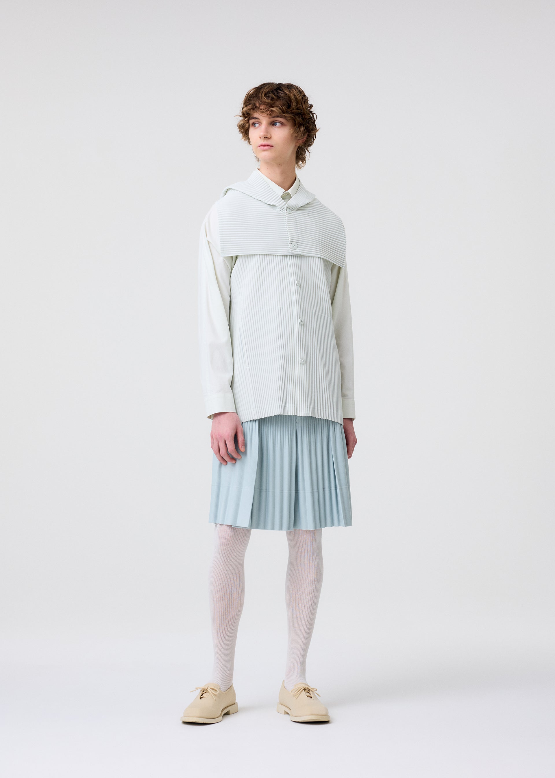 Zoomed view A person with curly hair wears a light, oversized button-up shirt and shoulder covering, paired with a blue pleated skirt from HOMME PLISSÉ ISSEY MIYAKE's MONTHLY COLORS : JUNE collection, white tights, and beige shoes against a white background.