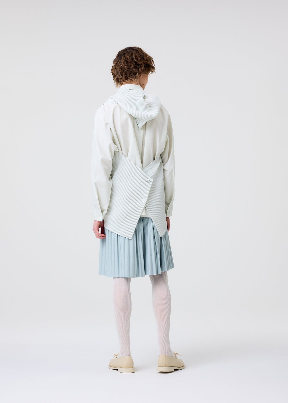 A person with short curly hair models HOMME PLISSÉ ISSEY MIYAKE's MONTHLY COLORS : JUNE, featuring lightweight pleated skirt-style shorts, white tights, cream shoes, and a unique structured white top with crossed pleats, on a plain white background. view 8 of 9