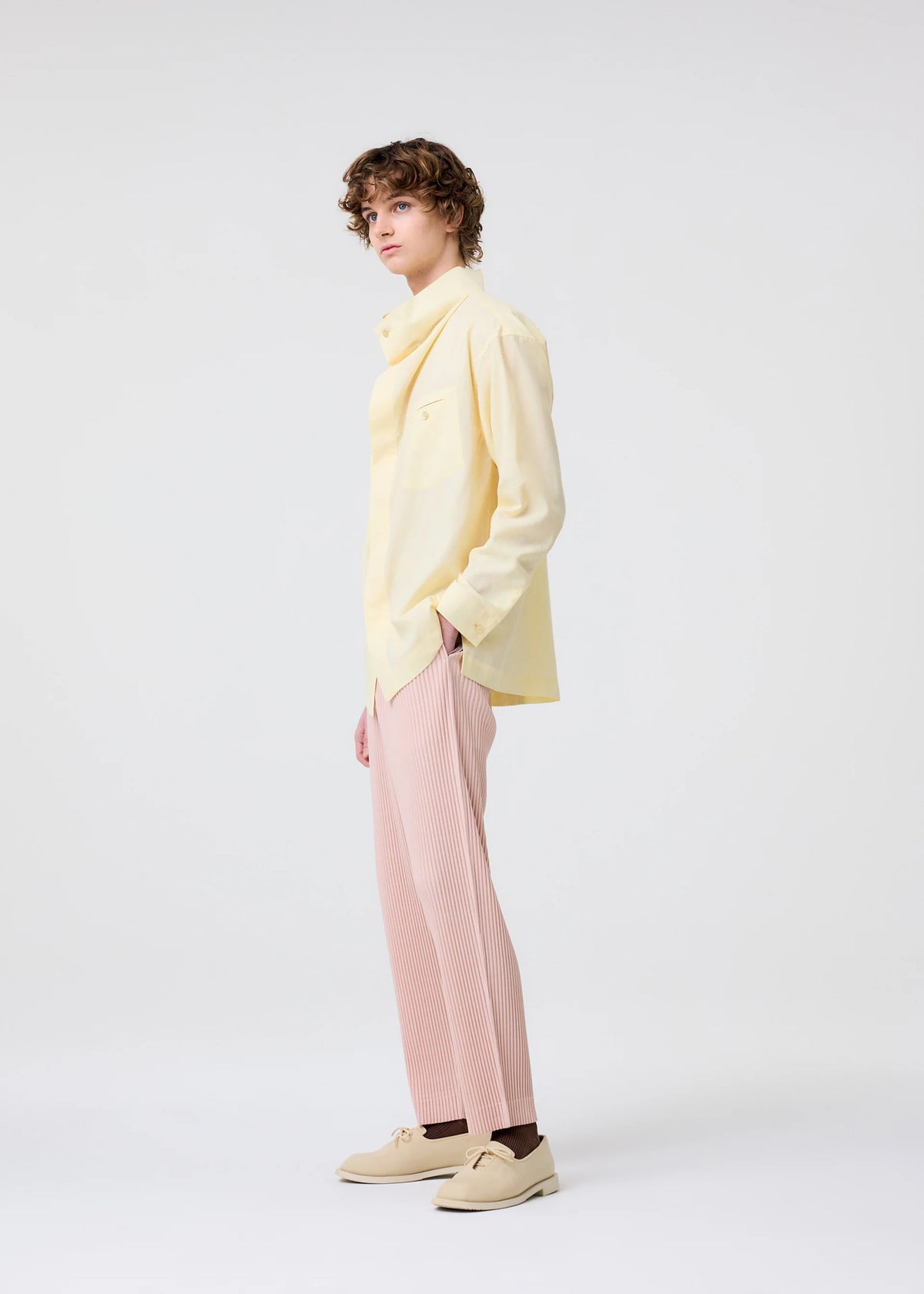 A person with short curly hair stands sideways against a light background, wearing a pale yellow button-up shirt, HOMME PLISSÉ ISSEY MIYAKE COLOR PLEATS 2 pants in pleated jersey, beige shoes, and has one hand in their pocket. view 5 of 6