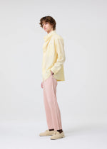 A person with short curly hair stands sideways against a light background, wearing a pale yellow button-up shirt, HOMME PLISSÉ ISSEY MIYAKE COLOR PLEATS 2 pants in pleated jersey, beige shoes, and has one hand in their pocket.