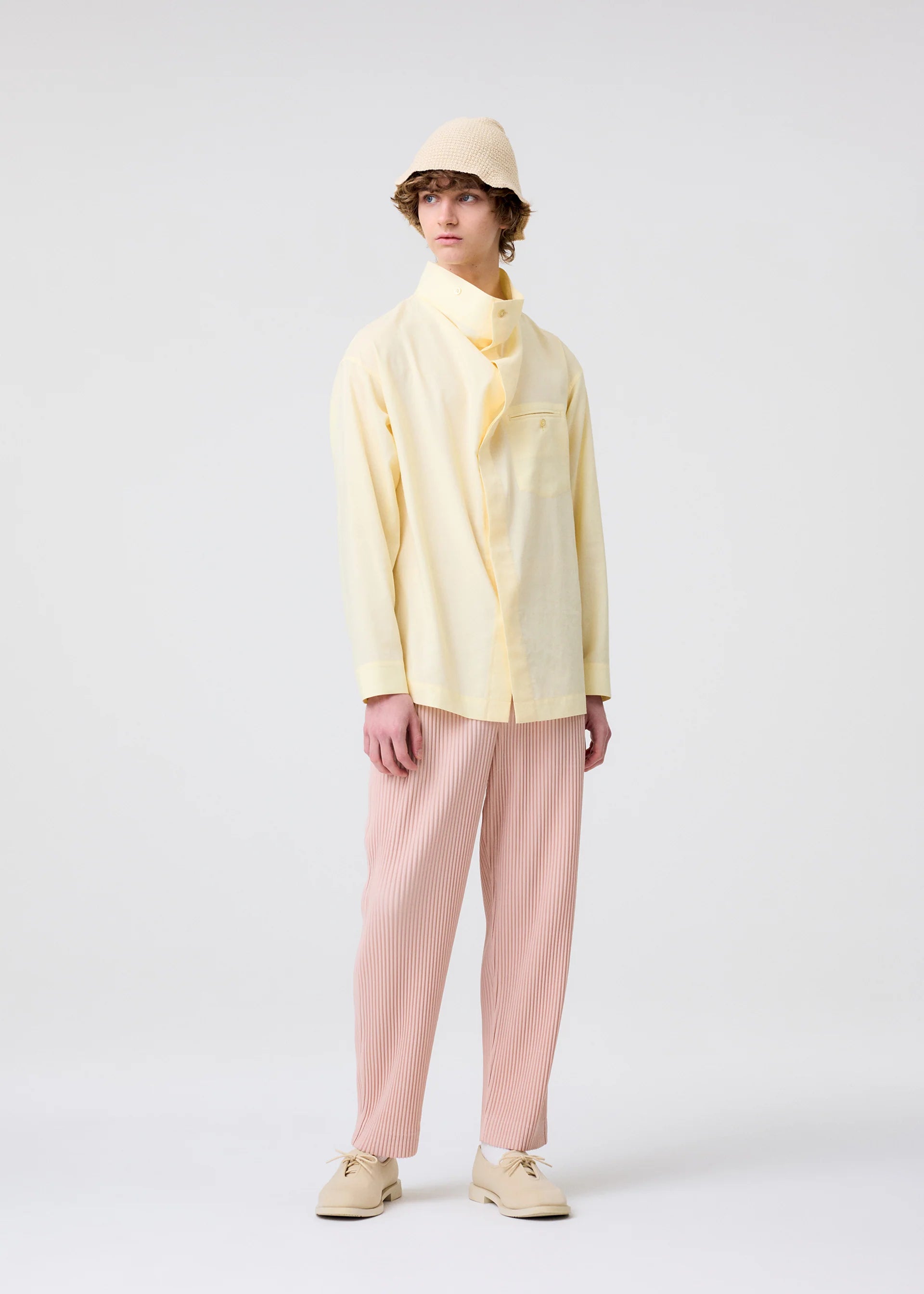 Zoomed view A person with short, curly hair and a neutral expression stands against a plain background wearing a pale yellow shirt, HOMME PLISSÉ ISSEY MIYAKE COLOR PLEATS 2 light pink pleated jersey pants, cream shoes, and a beige bucket hat.