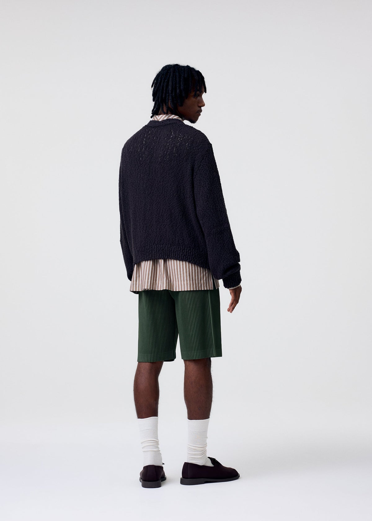 A person with short, twisted hair stands facing away, wearing a dark textured sweater over a long striped shirt, green COLOR PLEATS 2 shorts from HOMME PLISSÉ ISSEY MIYAKE, white socks, and dark shoes against a plain light background. view 5 of 5