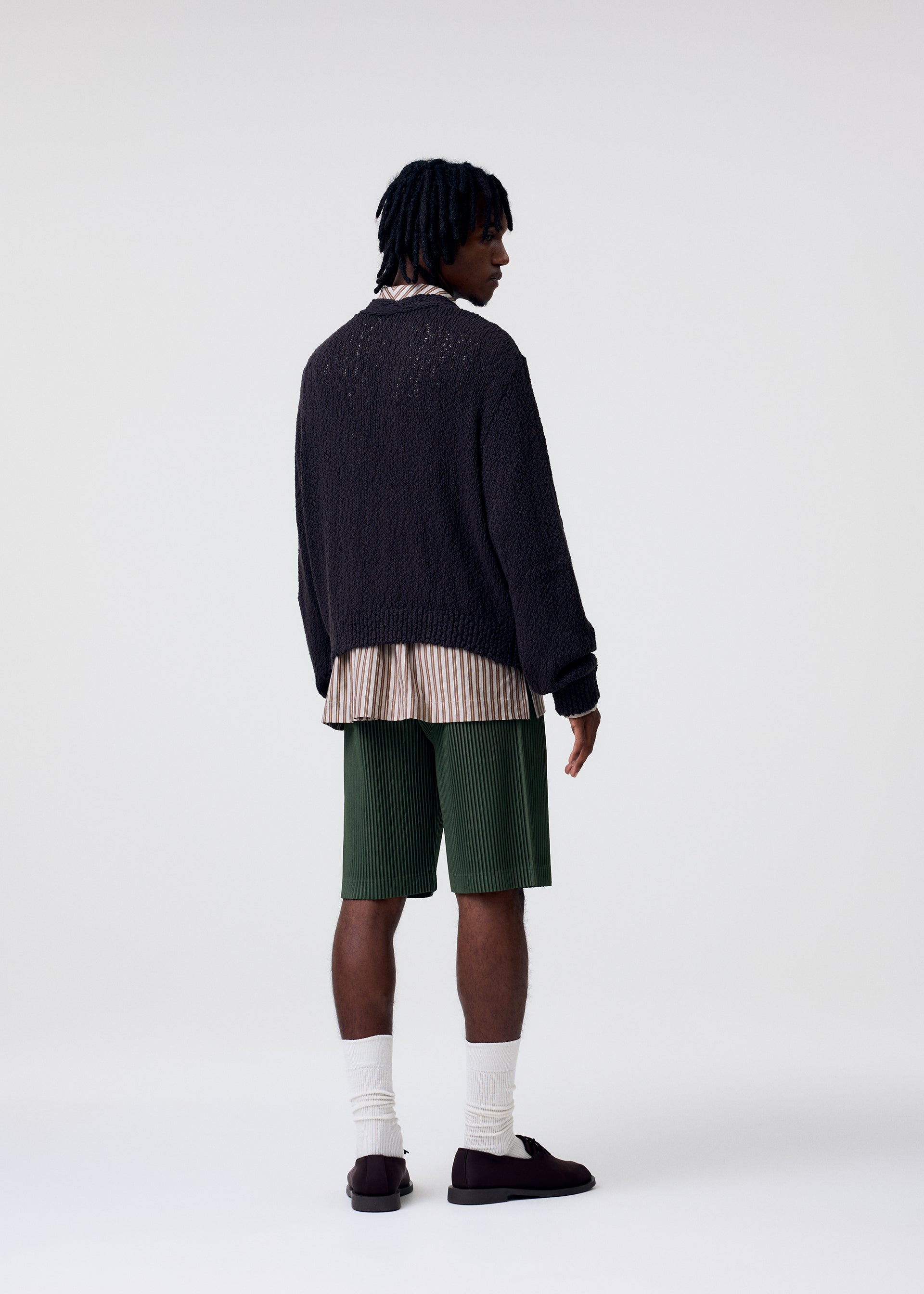 Zoomed view A person with short, twisted hair stands facing away, wearing a dark textured sweater over a long striped shirt, green COLOR PLEATS 2 shorts from HOMME PLISSÉ ISSEY MIYAKE, white socks, and dark shoes against a plain light background.