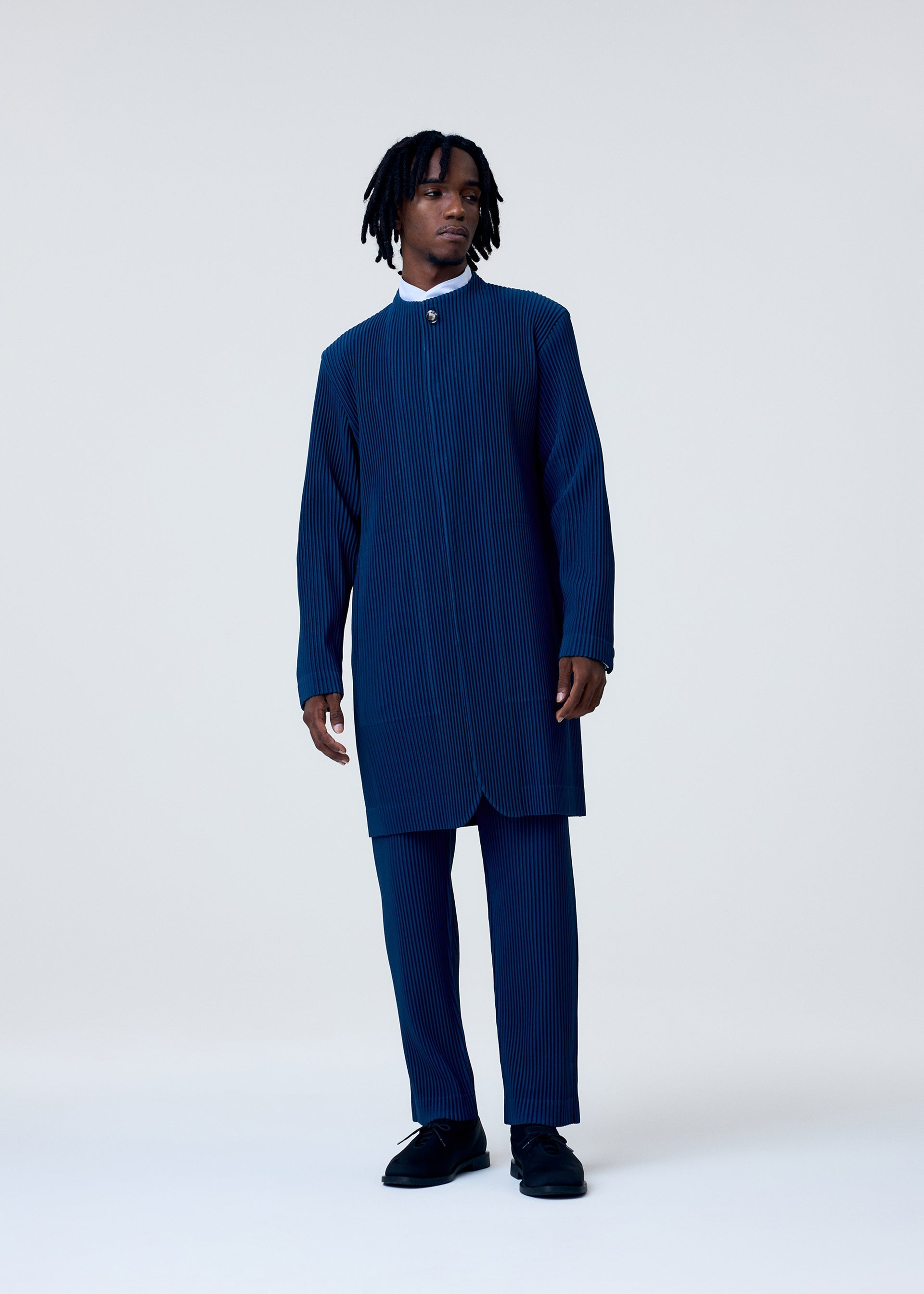 Zoomed view Wearing the TAILORED PLEATS 1 by HOMME PLISSÉ ISSEY MIYAKE, a person with medium-length dreadlocks pairs a blue pinstriped long coat with straight pants and black shoes, creating an elegant I-line silhouette against a plain light background.