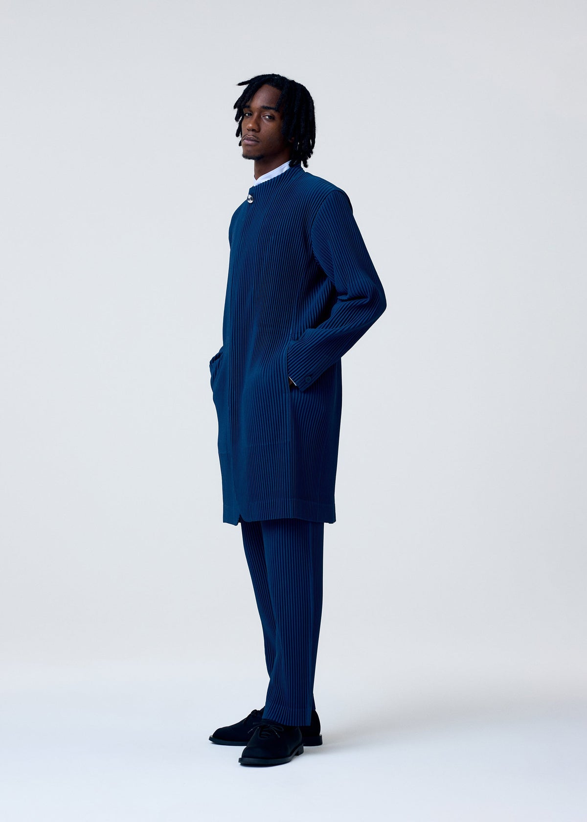 A man stands in profile against a light background, wearing HOMME PLISSÉ ISSEY MIYAKE's TAILORED PLEATS 1: a matching navy blue pinstriped I-line coat and straight pants, hands in pockets, completed with black shoes. view 5 of 6