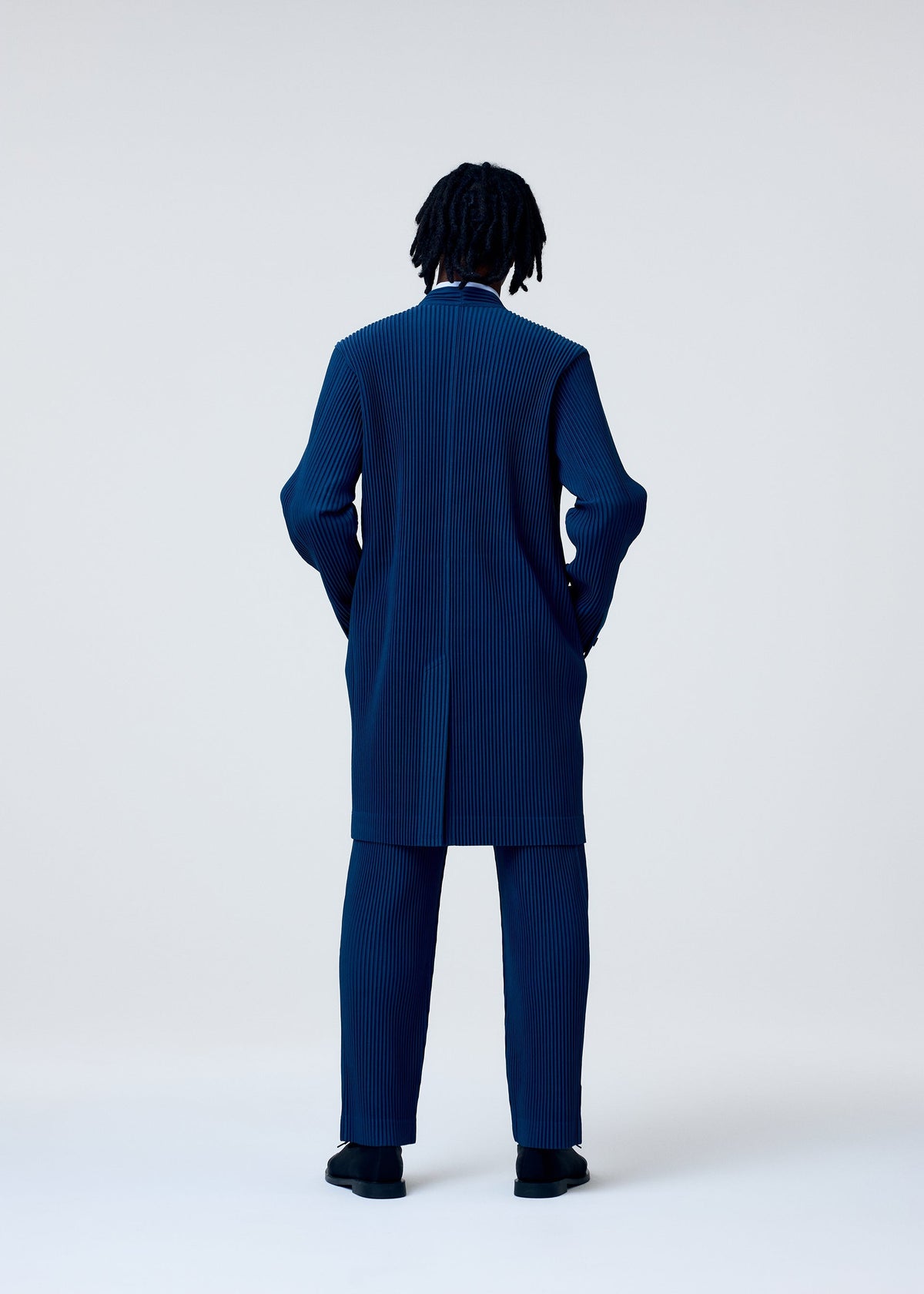 A person with short dreadlocks stands with their back to the camera, wearing the TAILORED PLEATS 1 coat and straight pants by HOMME PLISSÉ ISSEY MIYAKE in a minimalist studio setting. view 6 of 6