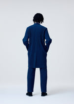 A person with short dreadlocks stands with their back to the camera, wearing the TAILORED PLEATS 1 coat and straight pants by HOMME PLISSÉ ISSEY MIYAKE in a minimalist studio setting.