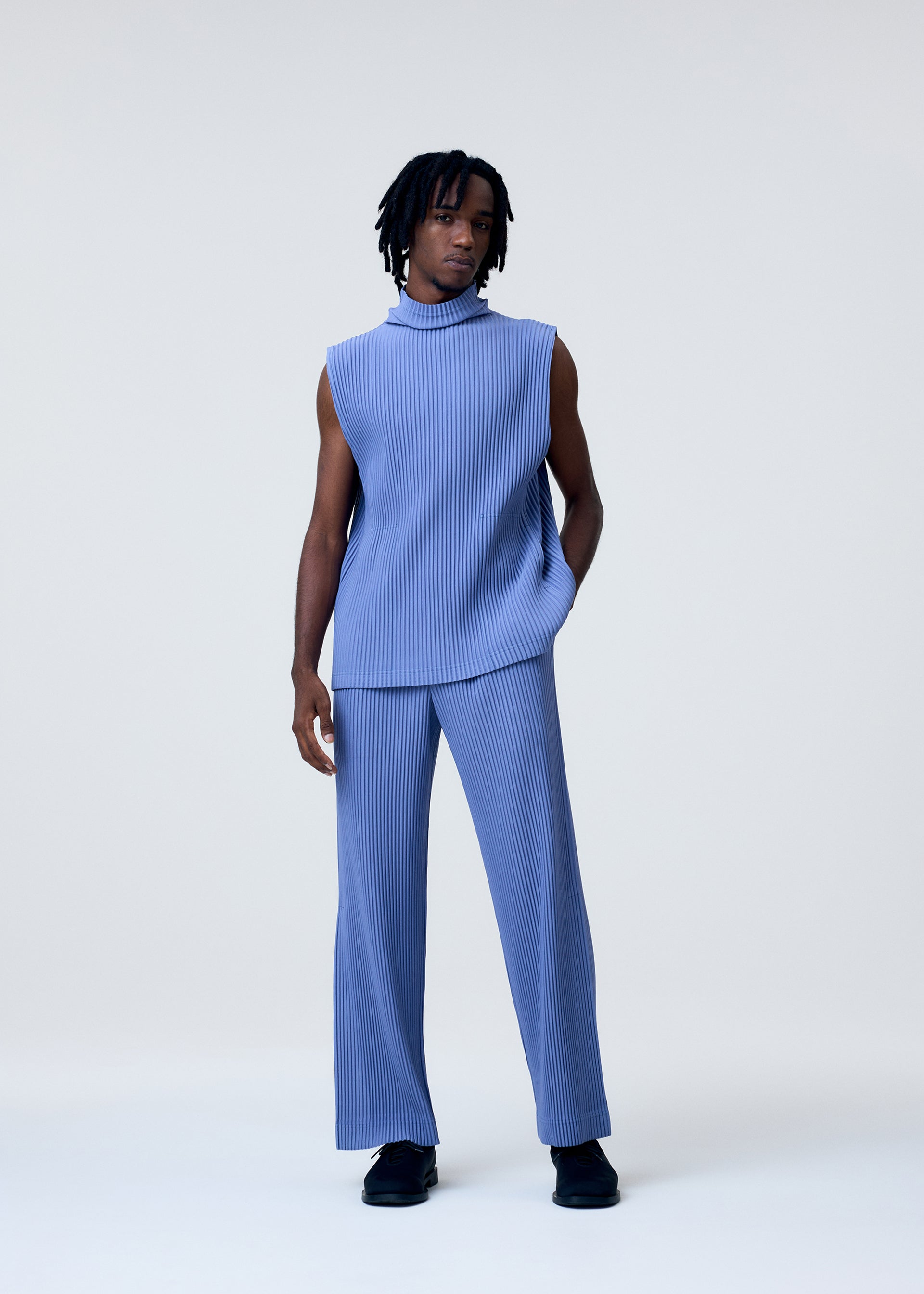 Zoomed view A person stands against a plain light background wearing the TAILORED PLEATS 1 set by HOMME PLISSÉ ISSEY MIYAKE, featuring a sleeveless, high-neck, ribbed blue top and matching flared pants, with black shoes and one hand in their pocket.