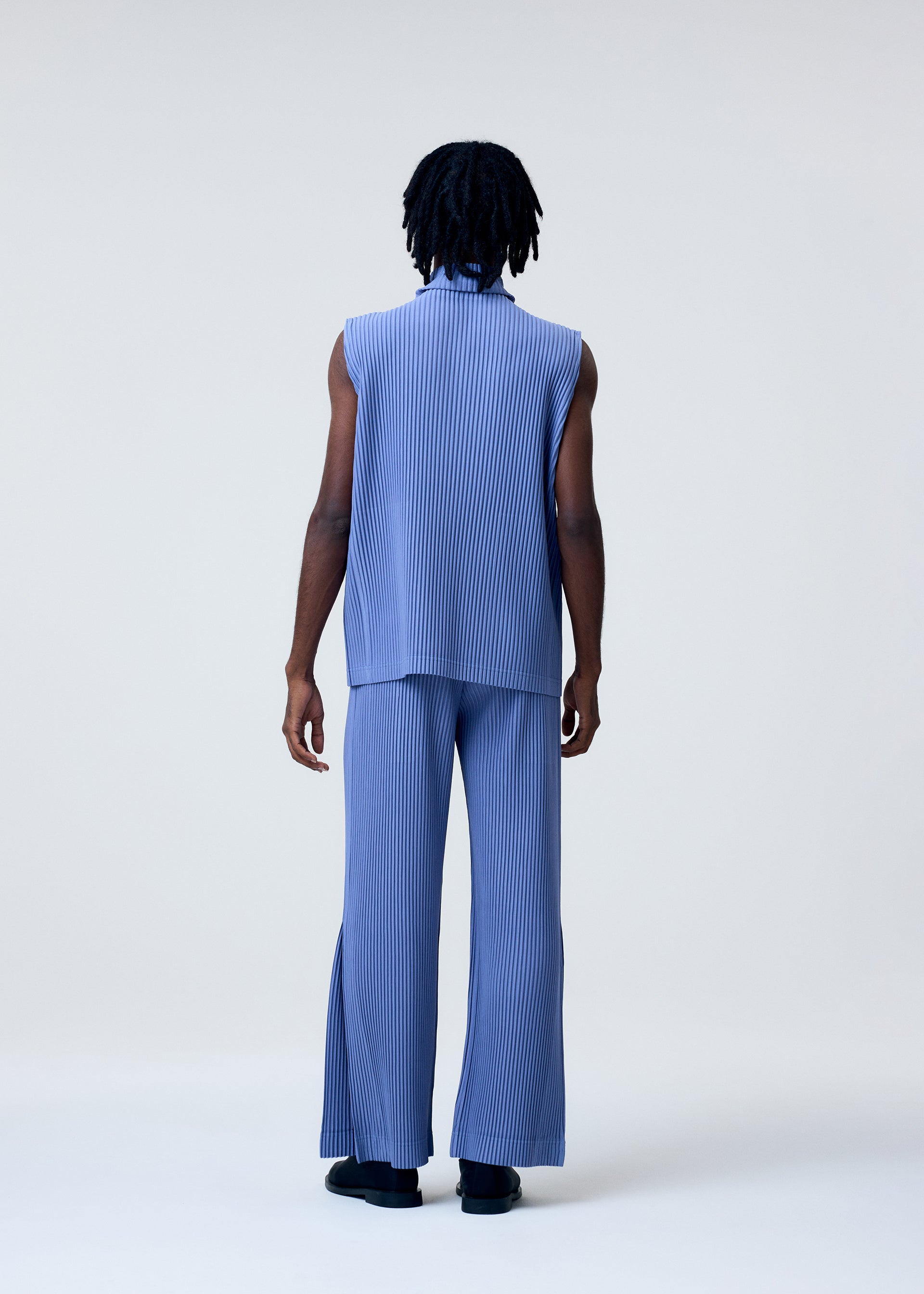 Zoomed view Someone with medium-dark skin and shoulder-length twisted hair faces away in a studio, wearing the HOMME PLISSÉ ISSEY MIYAKE TAILORED PLEATS 1: a loose, sleeveless light blue pleated top and matching wide-legged flared pants with dark shoes.