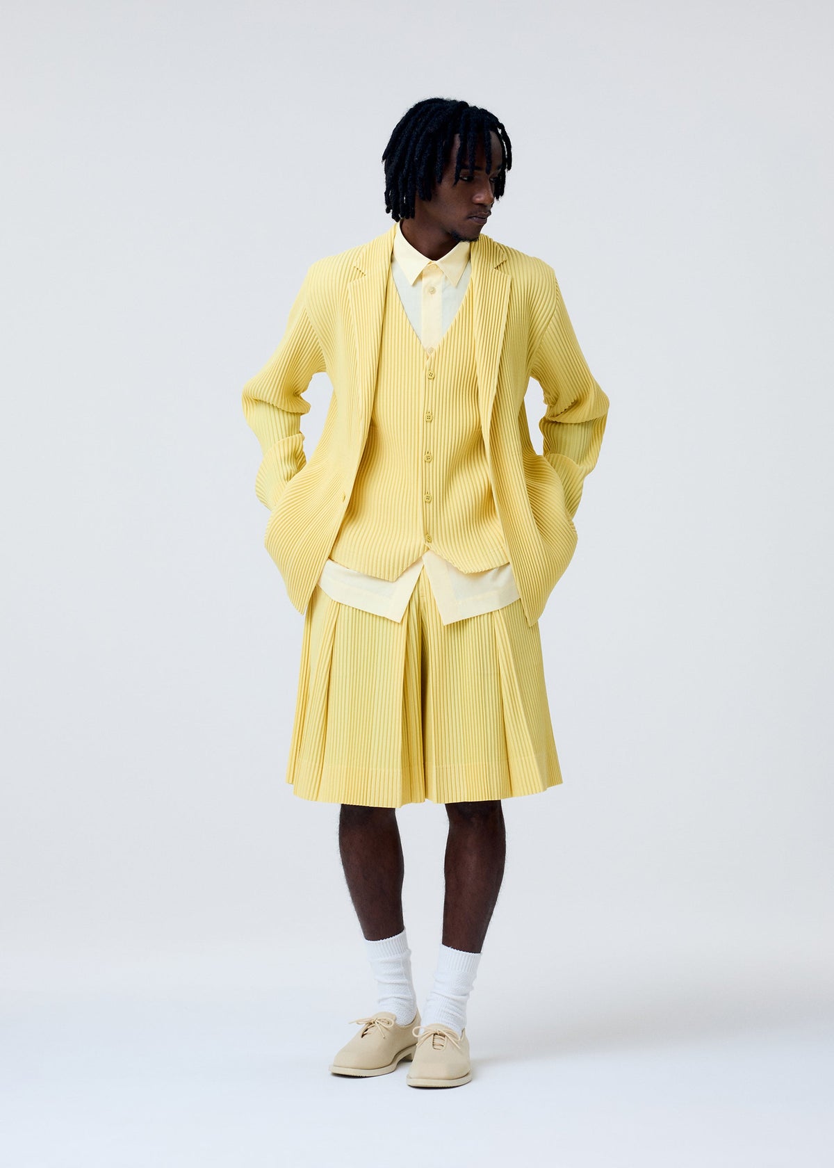 A person wears the HOMME PLISSÉ ISSEY MIYAKE TAILORED PLEATS 2: a coordinated yellow set with pleated blazer, vest, hakama-style shorts, white shirt and socks, and beige shoes, standing against a plain light background. view 8 of 9