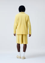 A person with short dreadlocks stands facing away against a plain light background, wearing HOMME PLISSÉ ISSEY MIYAKE’s TAILORED PLEATS 2—pleated yellow hakama-style shorts—with white socks and light-colored shoes.