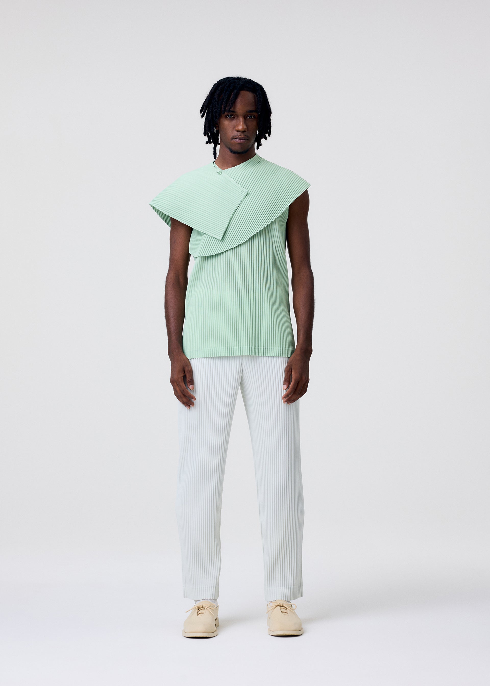 Zoomed view A person wears a sleeveless, mint green TAILORED PLEATS 3 top by HOMME PLISSÉ ISSEY MIYAKE with an asymmetrical flap over one shoulder, light straight-leg trousers, and cream shoes against a plain background.
