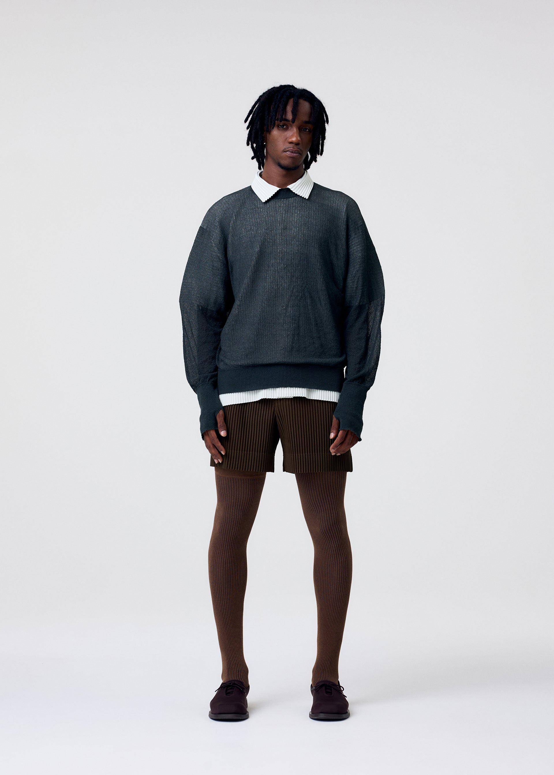 Zoomed view A person with short dreadlocks wears HOMME PLISSÉ ISSEY MIYAKE TAILORED PLEATS 3 shorts, a dark textured sweater over a white collared shirt, brown ribbed tights, and dark shoes, standing against a light background.