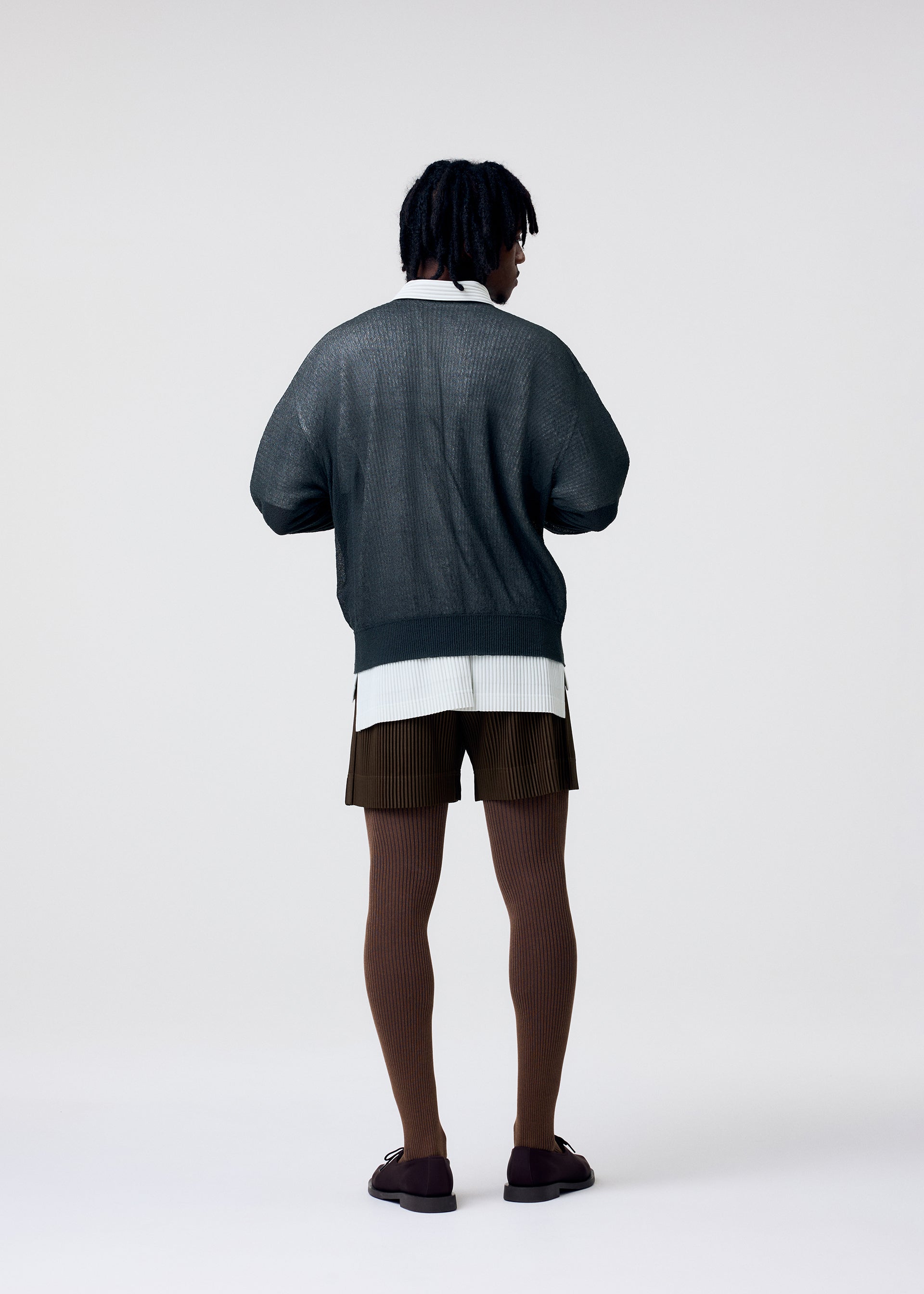 Zoomed view Wearing HOMME PLISSÉ ISSEY MIYAKE TAILORED PLEATS 3 brown pleated shorts, a dark loose sweater layered over a white shirt, brown ribbed tights, and dark shoes, a person stands facing away against a plain light background.
