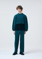A person with short curly hair wears the HOMME PLISSÉ ISSEY MIYAKE COLOR PLEATS 1 teal and black pleated long-sleeve top and matching pants, creating a relaxed silhouette against a plain light gray background.