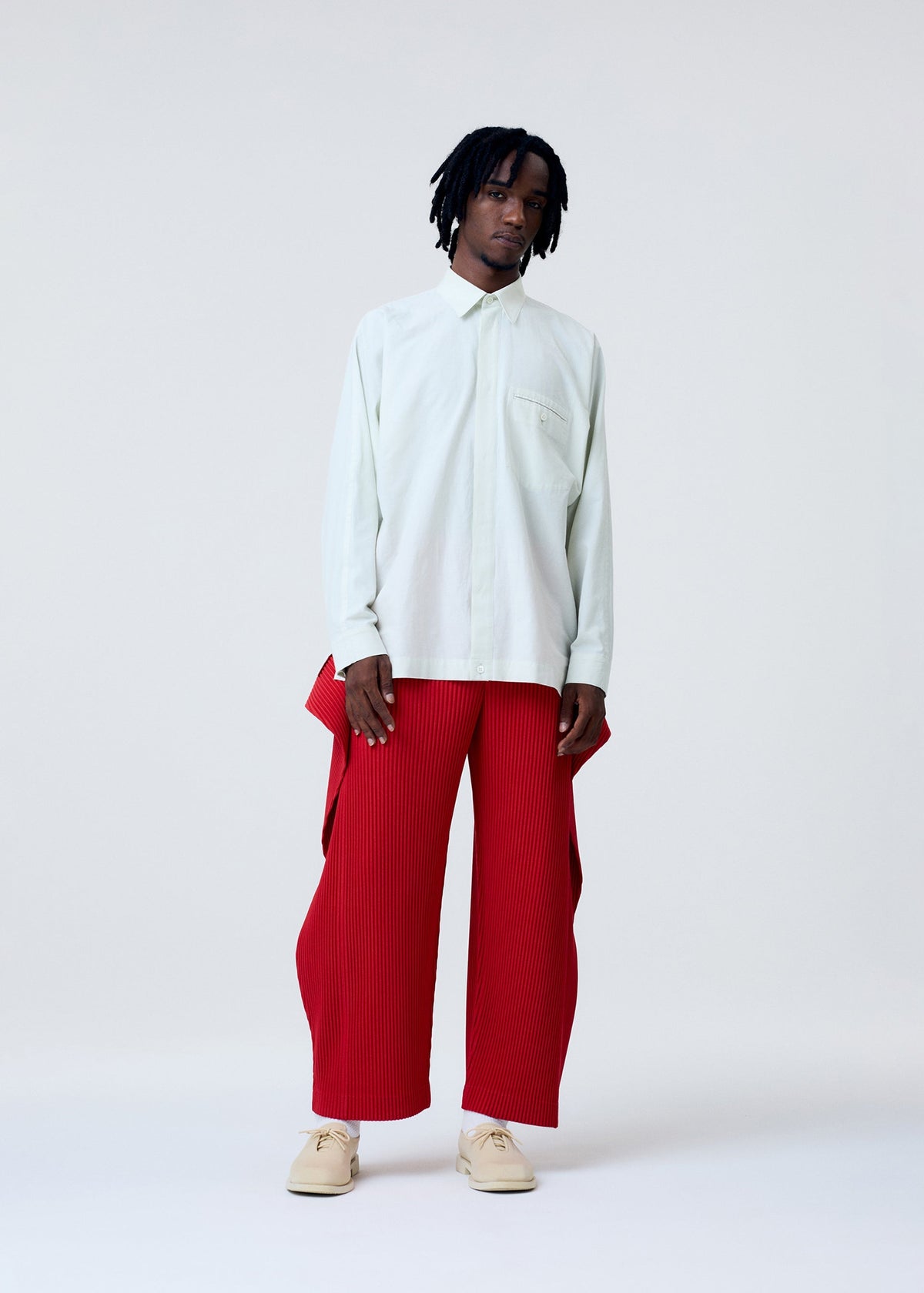 A person with short black dreadlocks wears the HOMME PLISSÉ ISSEY MIYAKE COVER AND FOLD: a loose light button-up shirt and bright red, wide-legged pleated pants with textured fabric, paired with beige shoes against a plain light background. view 6 of 8