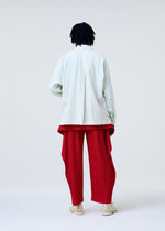 A person stands facing away, wearing the COVER AND FOLD bright red pleated pants with draped panels by HOMME PLISSÉ ISSEY MIYAKE, paired with a loose white long-sleeve shirt and cream-colored shoes against a plain light background.