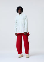 Against a white background, a person with short dreadlocks and a neutral expression wears the COVER AND FOLD shirt and bright red pleated pants from HOMME PLISSÉ ISSEY MIYAKE, styled with beige shoes.