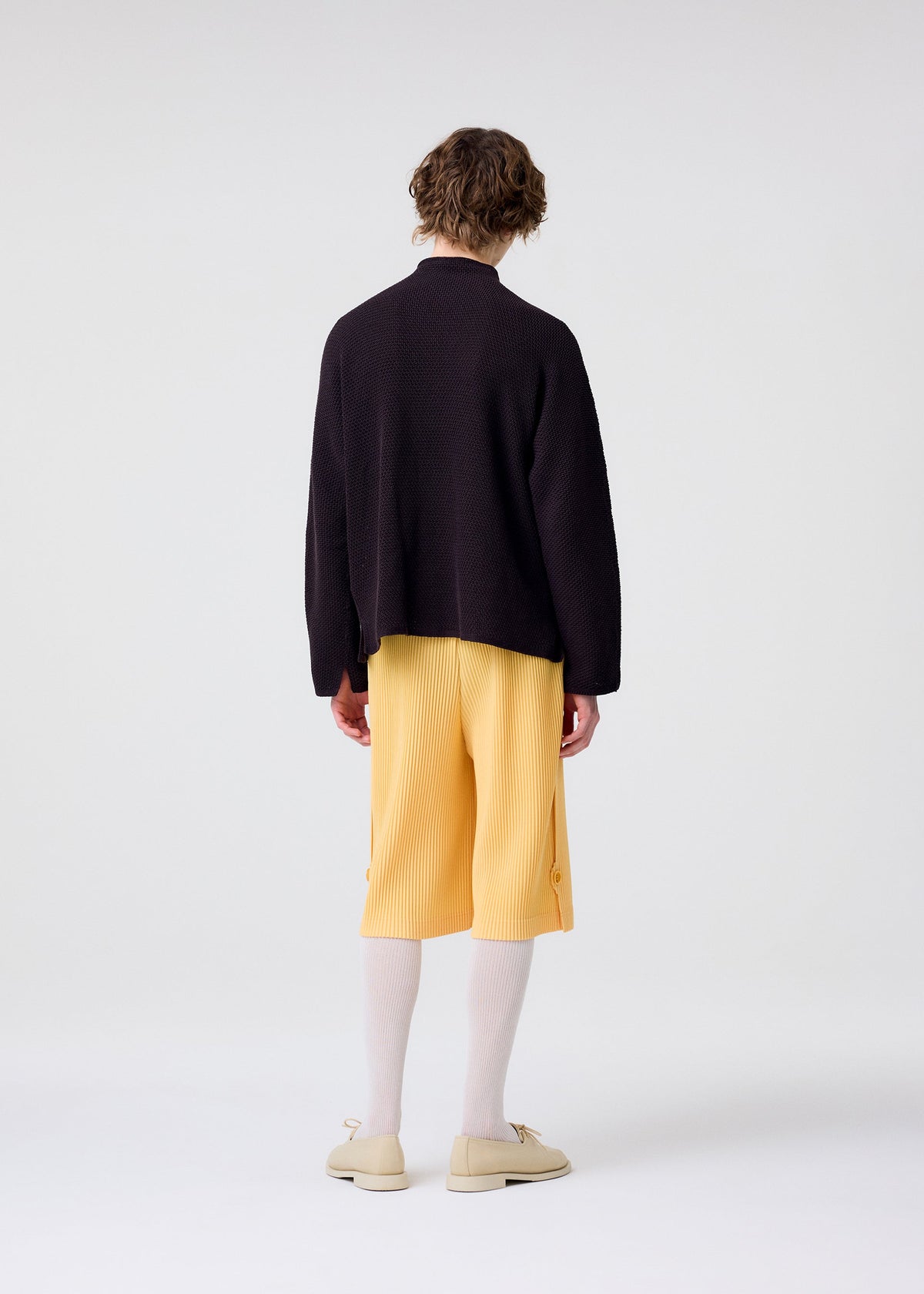 A person with short wavy hair, seen from behind, wears the HOMME PLISSÉ ISSEY MIYAKE WEAR OR WRAP loose dark sweater and yellow pleated shorts, with white knee-high socks and beige shoes, standing against a plain light background. view 8 of 9